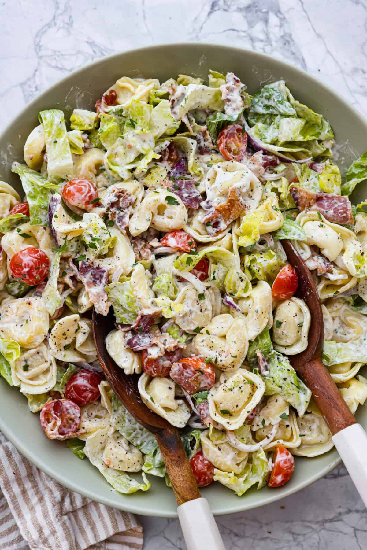 BLT Tortellini Pasta Salad is your favorite sandwich turned into a delicious, refreshing salad you can share with others. This salad is so easy to make and perfect for serving at any gathering. With loads of bacon, crisp Romaine lettuce, juicy tomatoes, and cheese-filled tortellini, it's sure to be a hit! Overhead shot of BLT tortellini pasta salad in large bowl with wooden spoon.