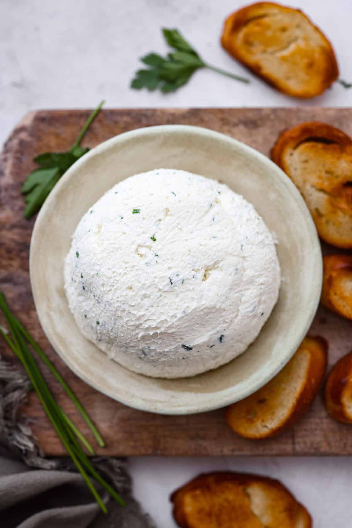 Boursin cheese is soft, creamy, and bursting with herbal flavor. Its texture is similar to cream cheese, making it incredibly versatile. It shines as a dip, enhances pasta dishes, complements meats, enriches sauces and soups, and so much more! Overhead shot of Boursin cheese ball on plate next to crostini's and chives.