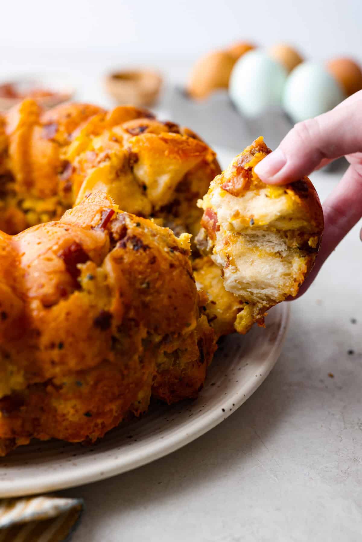 Rise and shine to Breakfast Monkey Bread with delicious savory flavors! It’s made with Grands biscuits, eggs, sausage, bacon, and cheese, all baked in a bundt pan. The result is crowd-pleasing pull-apart bread that everyone will devour! Side shot of someone pulling a piece of the savory breakfast monkey bread from the whole.