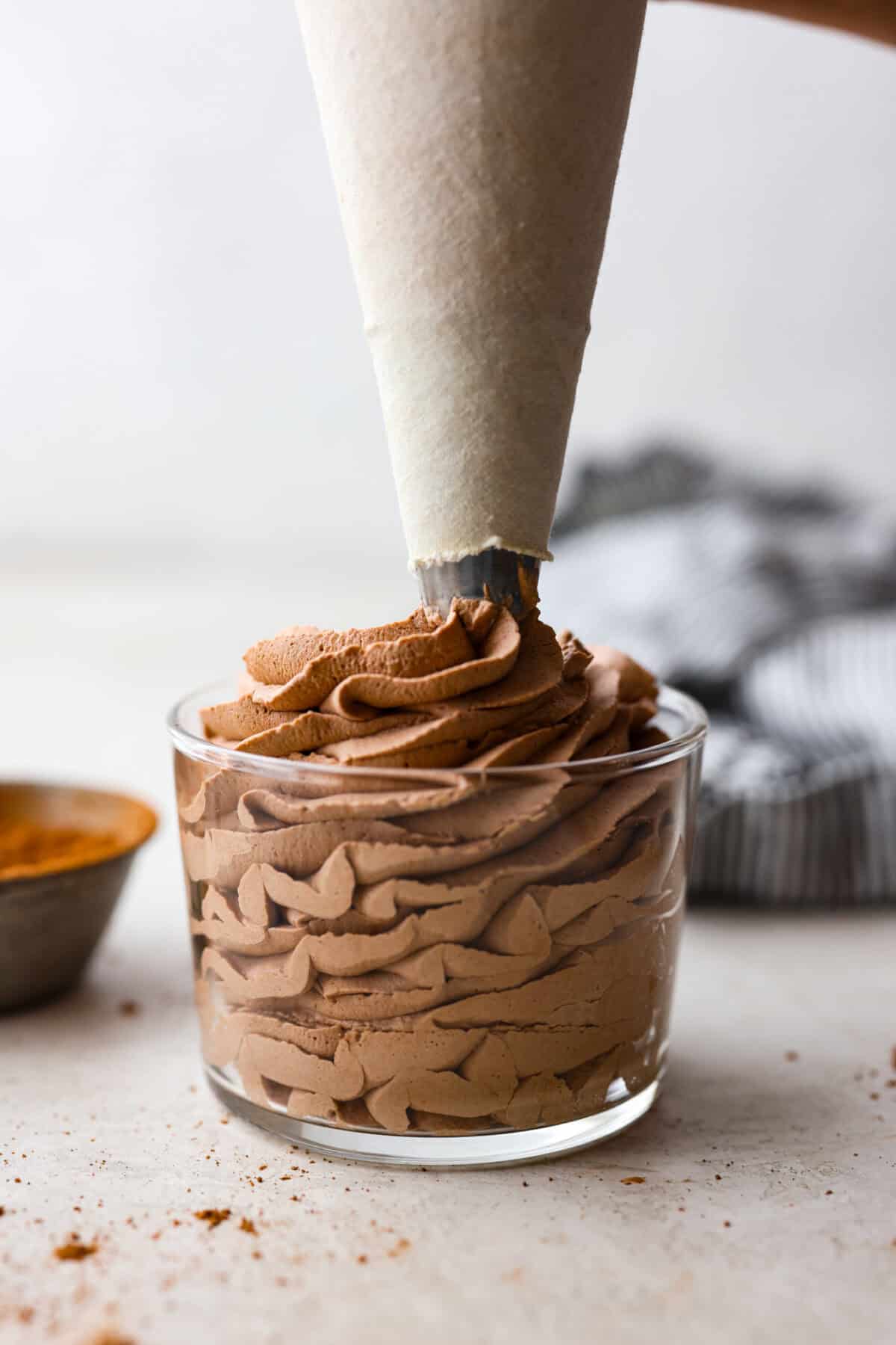 Fluffy Chocolate Whipped Cream is a wonderful combination of chocolate with fresh whipped cream! Itโs the perfect topping for fruit, waffles, pie, cupcakes and so much more! Side shot of someone piping chocolate whipped cream into a small glass container.