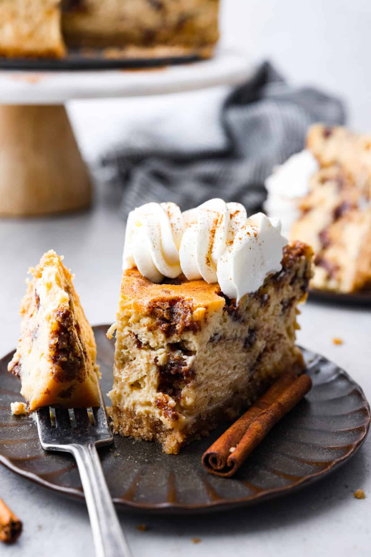 Rich and creamy Cinnamon Roll Cheesecake has the delicious flavor of a cinnamon roll in decadent cheesecake form. This irresistible dessert is made with a cinnamon graham cracker crust, a velvety cream cheese filling with spiced cinnamon throughout, and a classic whipped cream topping. Side shot of plated slice of cinnamon roll cheesecake with a bit on a fork.