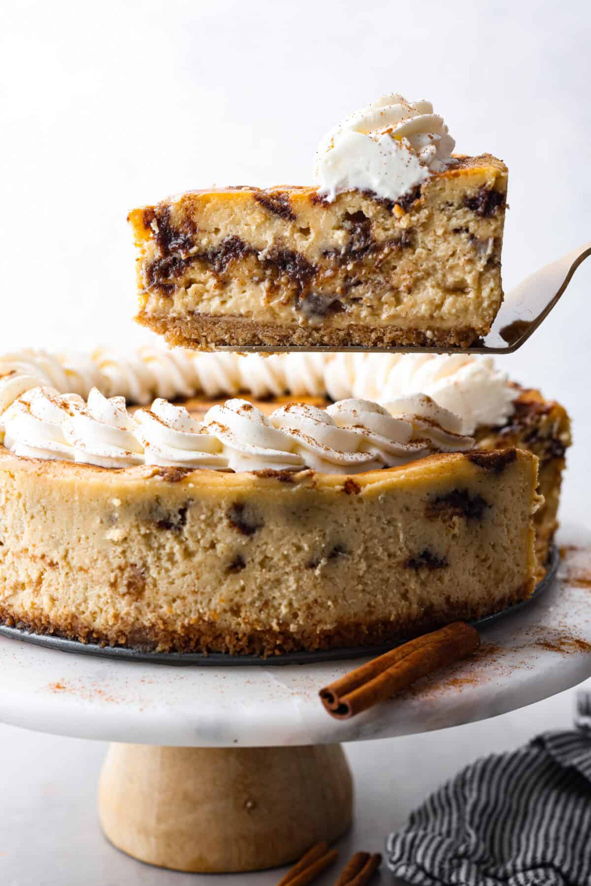 Rich and creamy Cinnamon Roll Cheesecake has the delicious flavor of a cinnamon roll in decadent cheesecake form. This irresistible dessert is made with a cinnamon graham cracker crust, a velvety cream cheese filling with spiced cinnamon throughout, and a classic whipped cream topping. Side shot of slice of cheesecake being lifted out of the whole cinnamon roll cheesecake.