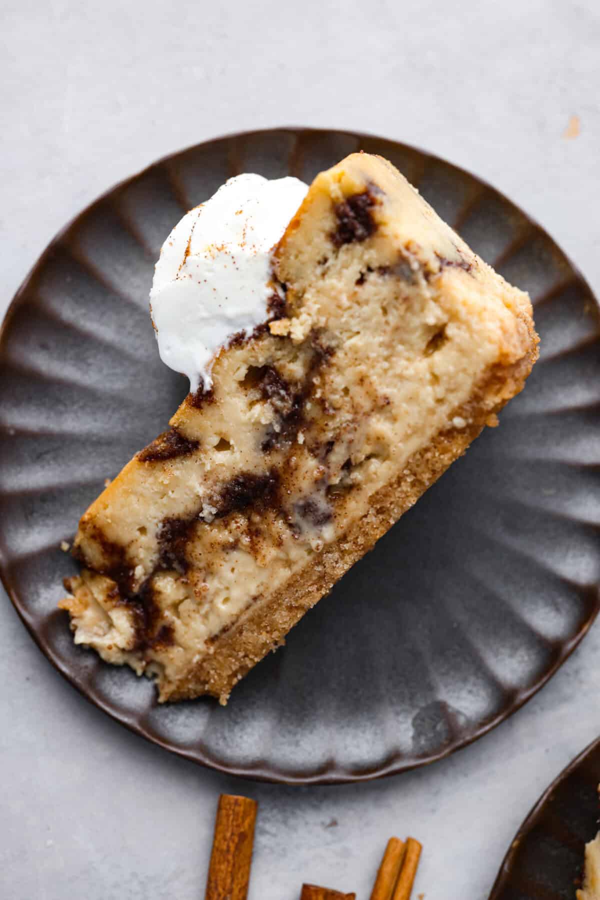 Rich and creamy Cinnamon Roll Cheesecake has the delicious flavor of a cinnamon roll in decadent cheesecake form. This irresistible dessert is made with a cinnamon graham cracker crust, a velvety cream cheese filling with spiced cinnamon throughout, and a classic whipped cream topping. Overhead shot of a slice of cinnamon roll cheesecake on a plate tipped on its side.