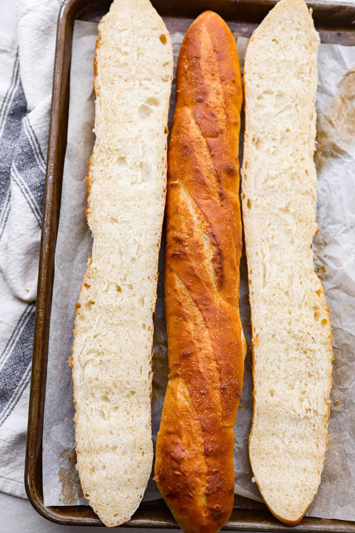 This French baguette recipe is super easy and will change your life! It has a perfect crusty outside and tender, soft inside, perfect for your bread cravings. Overhead shot of baked French baguette loaves on a baking sheet, one is cut in half the long way.