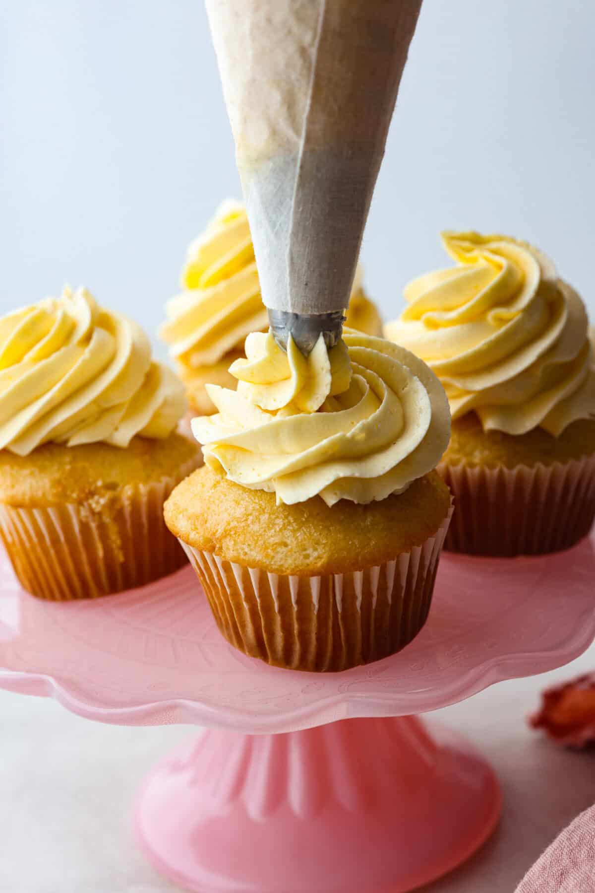 French buttercream is a luxurious, creamy frosting that will wow your guests and tantalize your taste buds. The elegant flavor comes from using egg yolks as the base and delicious, simple syrup for sweetness. Elevate your baking game with this unique recipe. Side shot of french buttercream being piped onto a cupcake that is on a pink cake stand with other cupcakes.