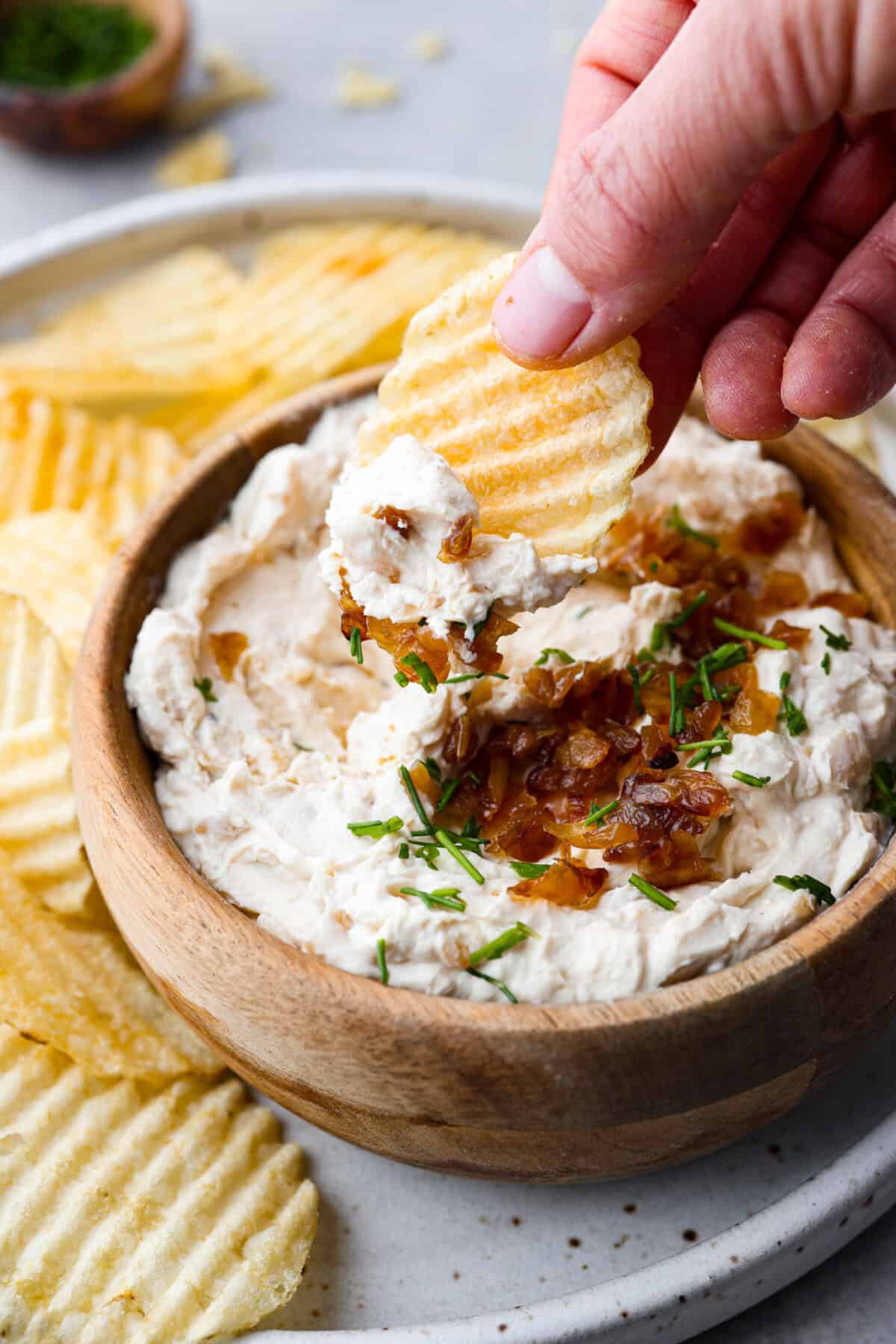 The Best French Onion Dip begins with caramelized onions that combine with cream cheese, sour cream, and seasonings. It creates a dip so addictive and creamy, it'll be gone before you know it! Close view of a chip dipping into bowl of French onion dip.