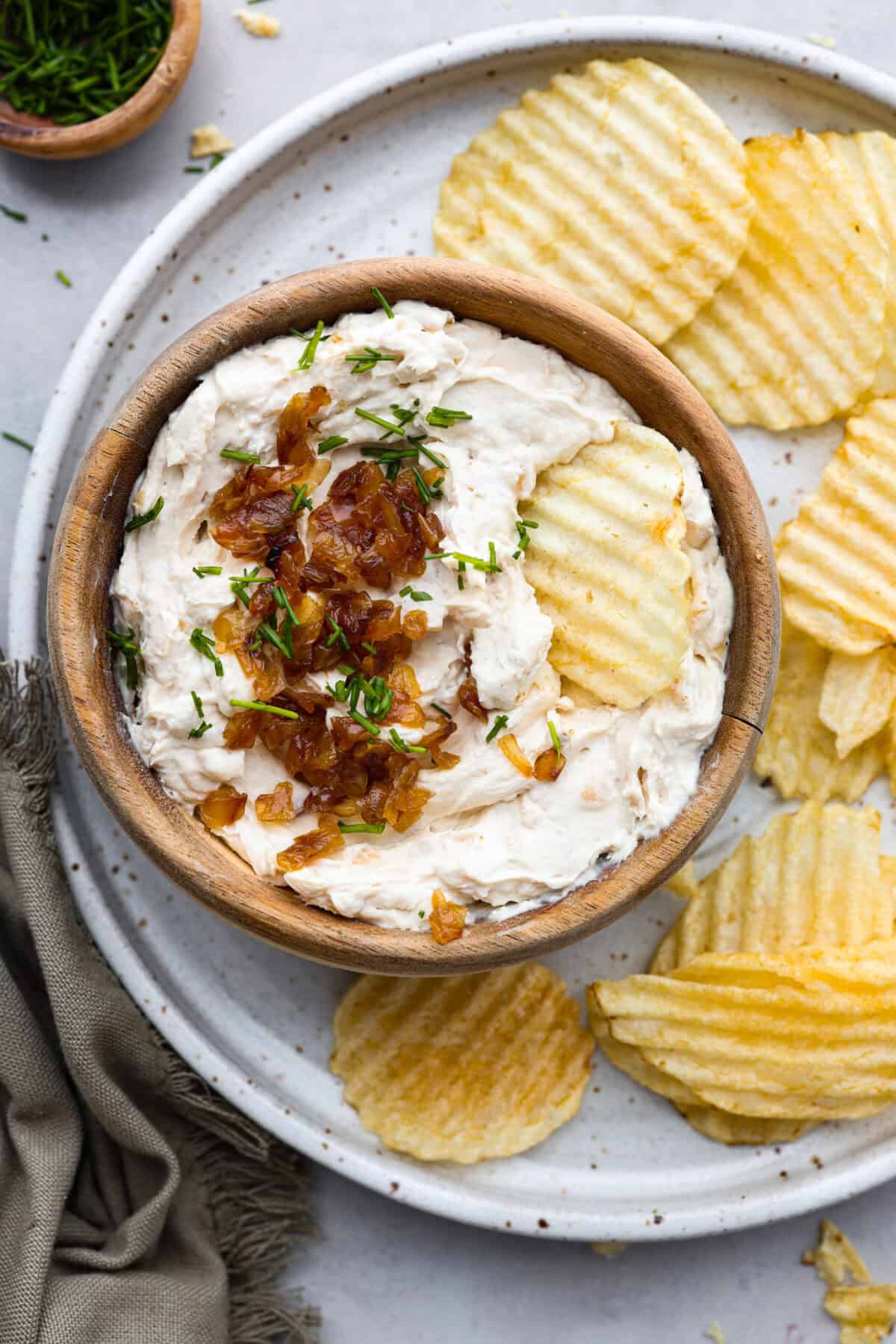 The Best French Onion Dip begins with caramelized onions that combine with cream cheese, sour cream, and seasonings. It creates a dip so addictive and creamy, it'll be gone before you know it! Top view of French onion dip in a wood bowl with chips on the side.