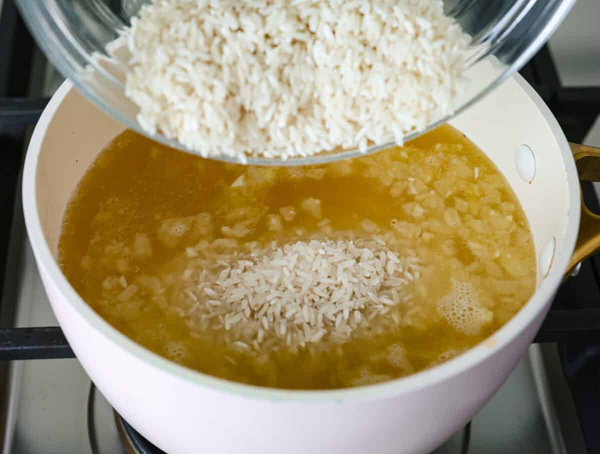 Garlic Butter Rice is the perfect side dish for dinner! A simpleย mix of butter, sautรฉed onion, garlic, and chicken brothย gives this rice tons of great flavor. It's easy to make and tastes amazing! Overhead shot of someone pouring rice into the broth, butter, onion and garlic mixture in the pot.