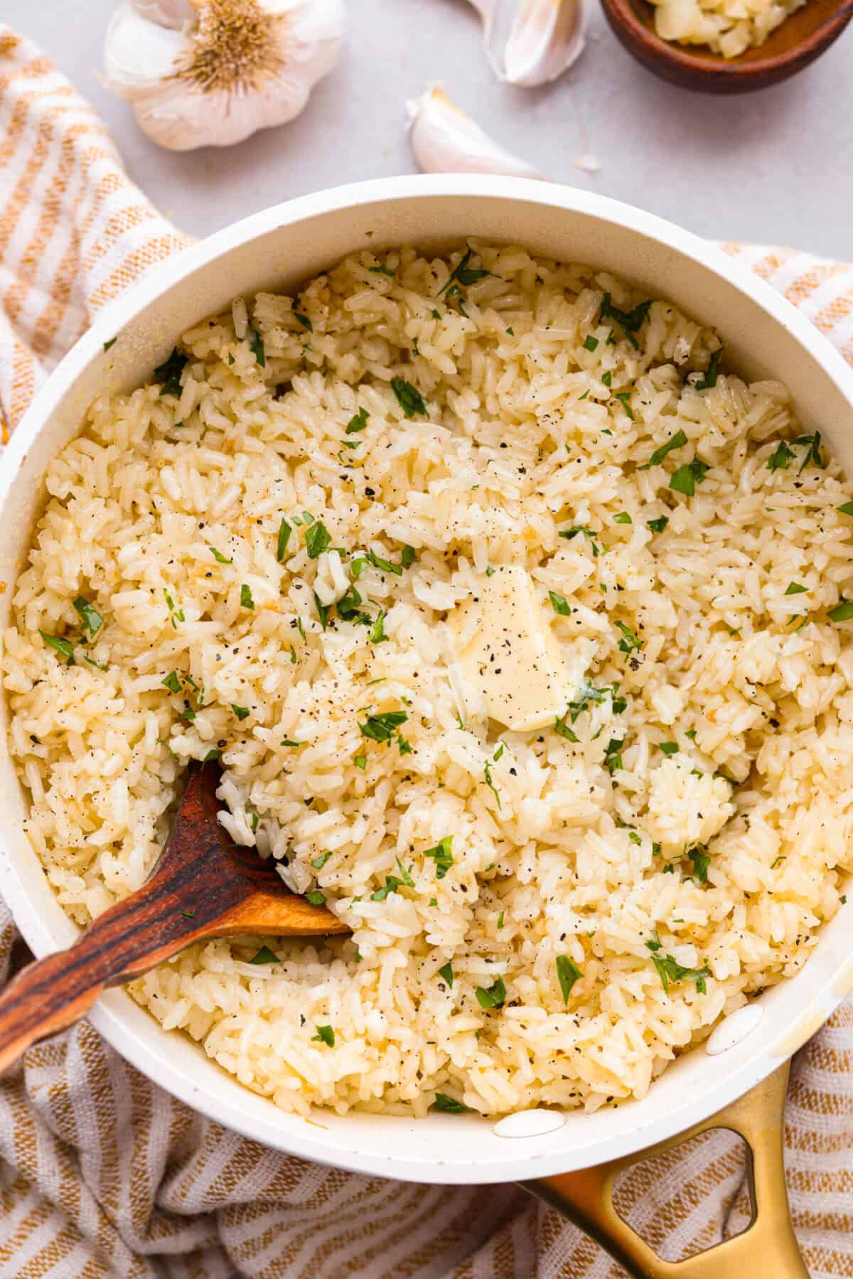 Garlic Butter Rice is the perfect side dish for dinner! A simpleย mix of butter, sautรฉed onion, garlic, and chicken brothย gives this rice tons of great flavor. It's easy to make and tastes amazing! Overhead shot of garlic butter rice in a pot with wooden spoon, garnished with parsley.