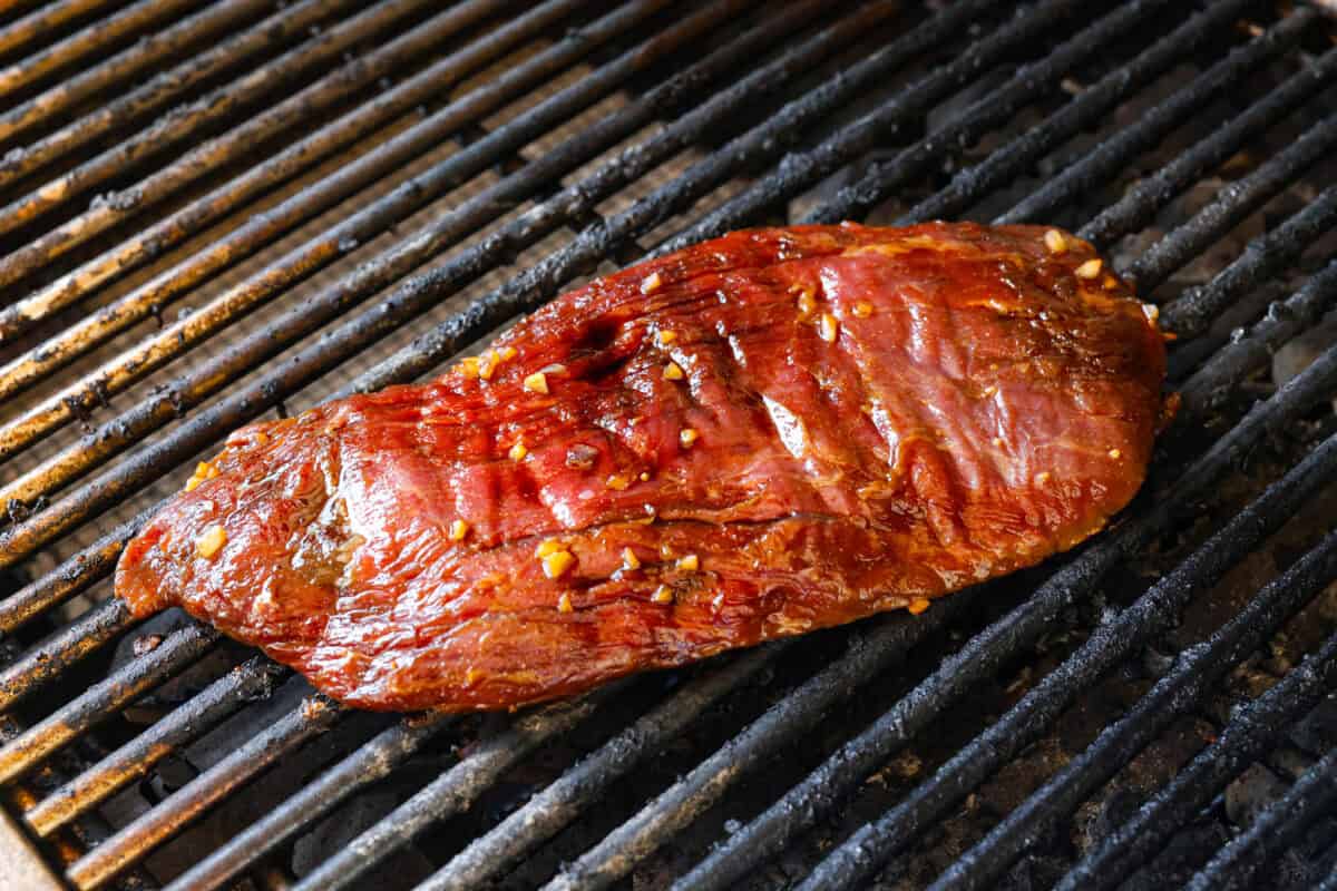 This grilled flank steak with Asian-inspired marinade features a savory blend of soy sauce, ginger, and garlic. The steak is seared to perfection on a hot grill, locking in all the delicious flavors. Best of all, itโs ready in less than 10 minutes! Angle shot of flank steak cooking on grill.
