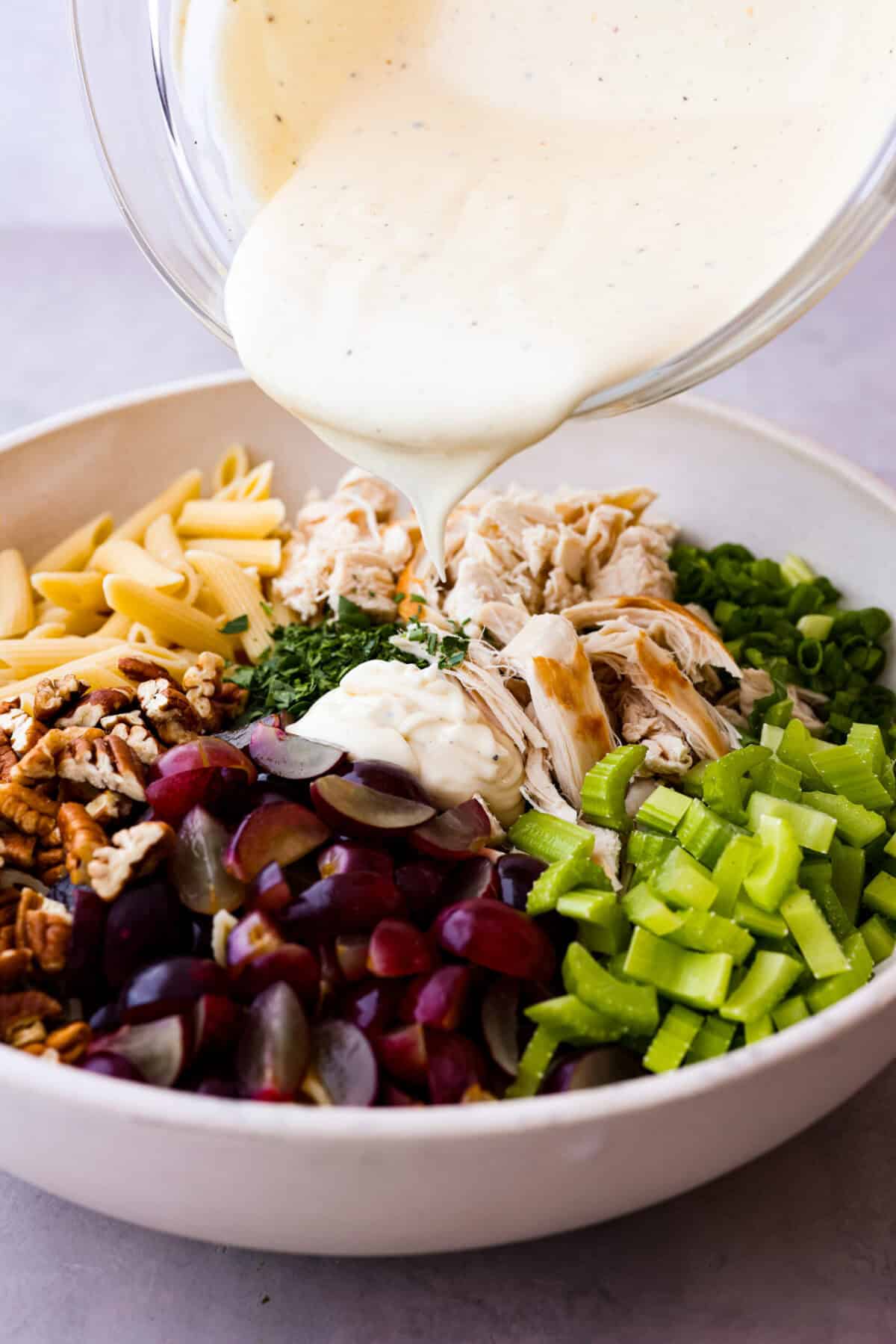 This healthy chicken pasta salad is a fun twist on the classic sandwich recipe. It's filled with penne pasta, tender chunks of chicken breast, crunchy pecans, all tossed in a creamy, sweet dressing. It's the perfect blend of flavors and textures. Side shot of dressing being poured over colorful pasta salad ingredients.