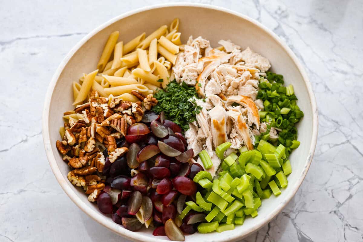 This healthy chicken pasta salad is a fun twist on the classic sandwich recipe. It's filled with penne pasta, tender chunks of chicken breast, crunchy pecans, all tossed in a creamy, sweet dressing. It's the perfect blend of flavors and textures. Overhead shot of bowl with pasta salad ingredients not mixed together in separate piles.