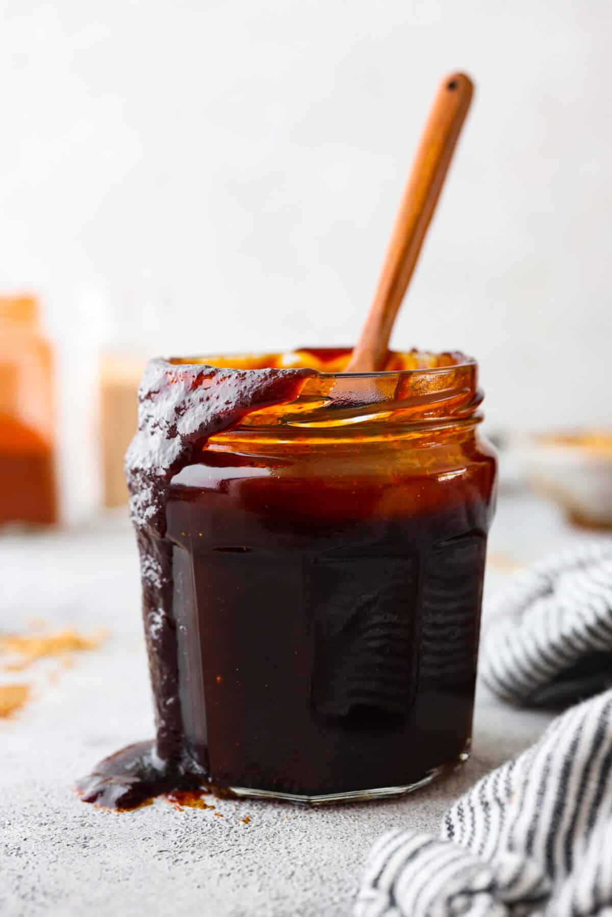 This Homemade BBQ Sauce recipe has the most amazing flavor combination and only takes minutes to make! It's sweet and tangy and has the perfect amount of smokiness that makes this BBQ sauce a family favorite! Close view of a jar of bbq sauce with a wooden spoon and sauce drizzling down the side of the jar.