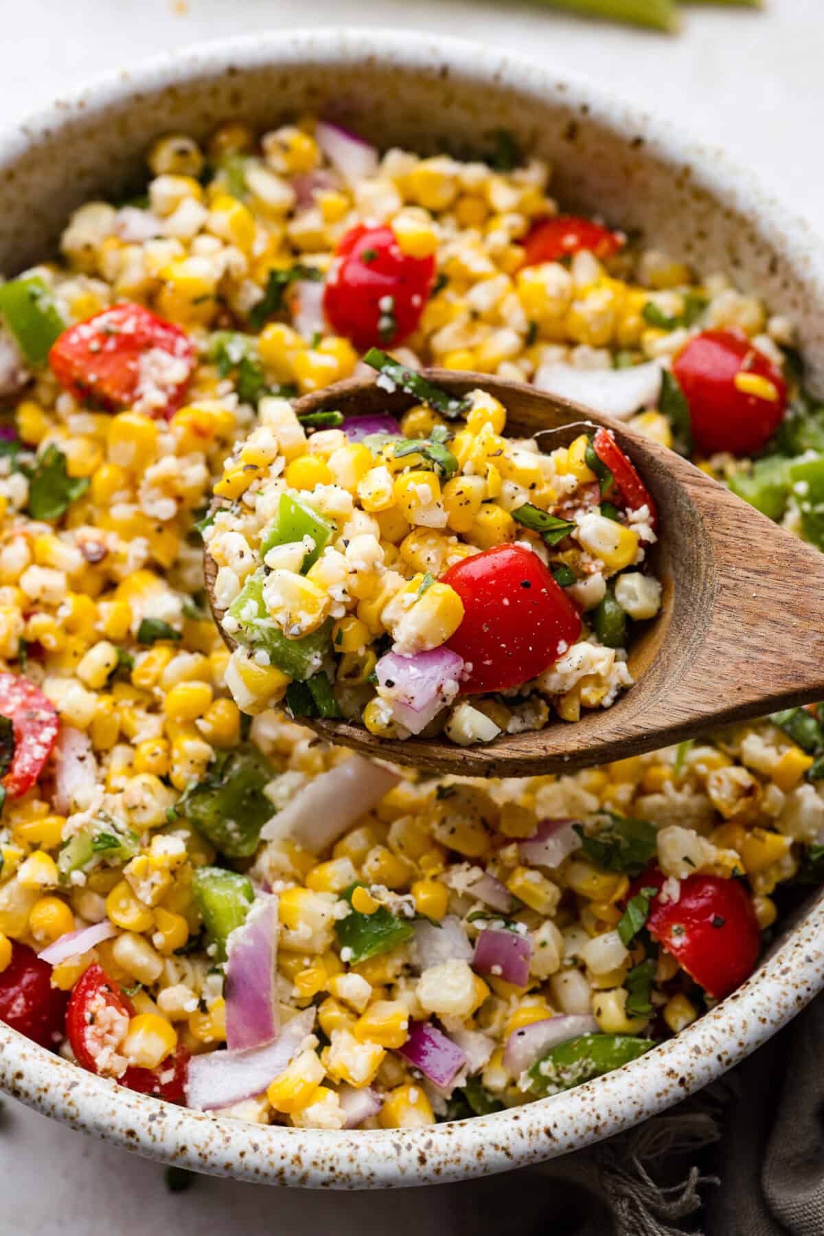 This Italian Corn Salad is bursting with fresh flavors from sweet corn, juicy grape tomatoes, crisp green bell pepper, and sharp parmesan cheese. Tossed in a zesty homemade Italian dressing, it’s perfect for summer or any time of the year. Angle shot of wooden spoon with scoop of Italian corn salad.