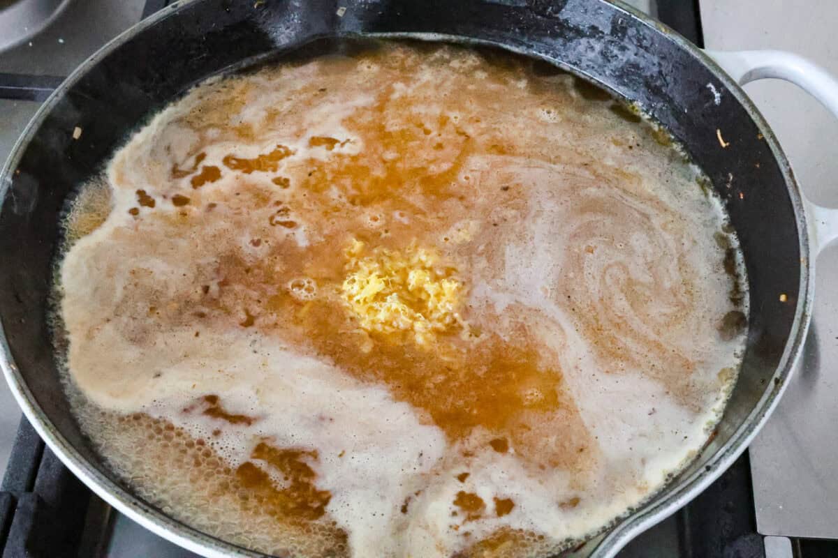 One-Pan Lemon Chicken Orzo is a comforting, well-rounded meal! Tender, juicy chicken pieces are joined with orzo pasta in a bright lemon sauce, resulting in melt-in-your-mouth deliciousness! Your whole family will adore this tasty dinner! Angle shot of orzo pasta with liquid and lemon zest in a pan.