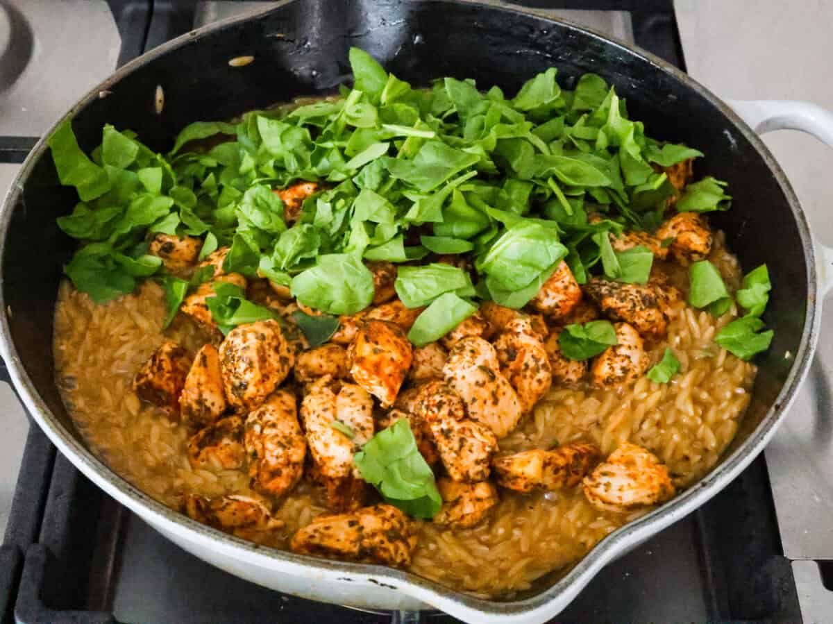One-Pan Lemon Chicken Orzo is a comforting, well-rounded meal! Tender, juicy chicken pieces are joined with orzo pasta in a bright lemon sauce, resulting in melt-in-your-mouth deliciousness! Your whole family will adore this tasty dinner! Angle shot of cooked chicken and baby spinach added to pan.