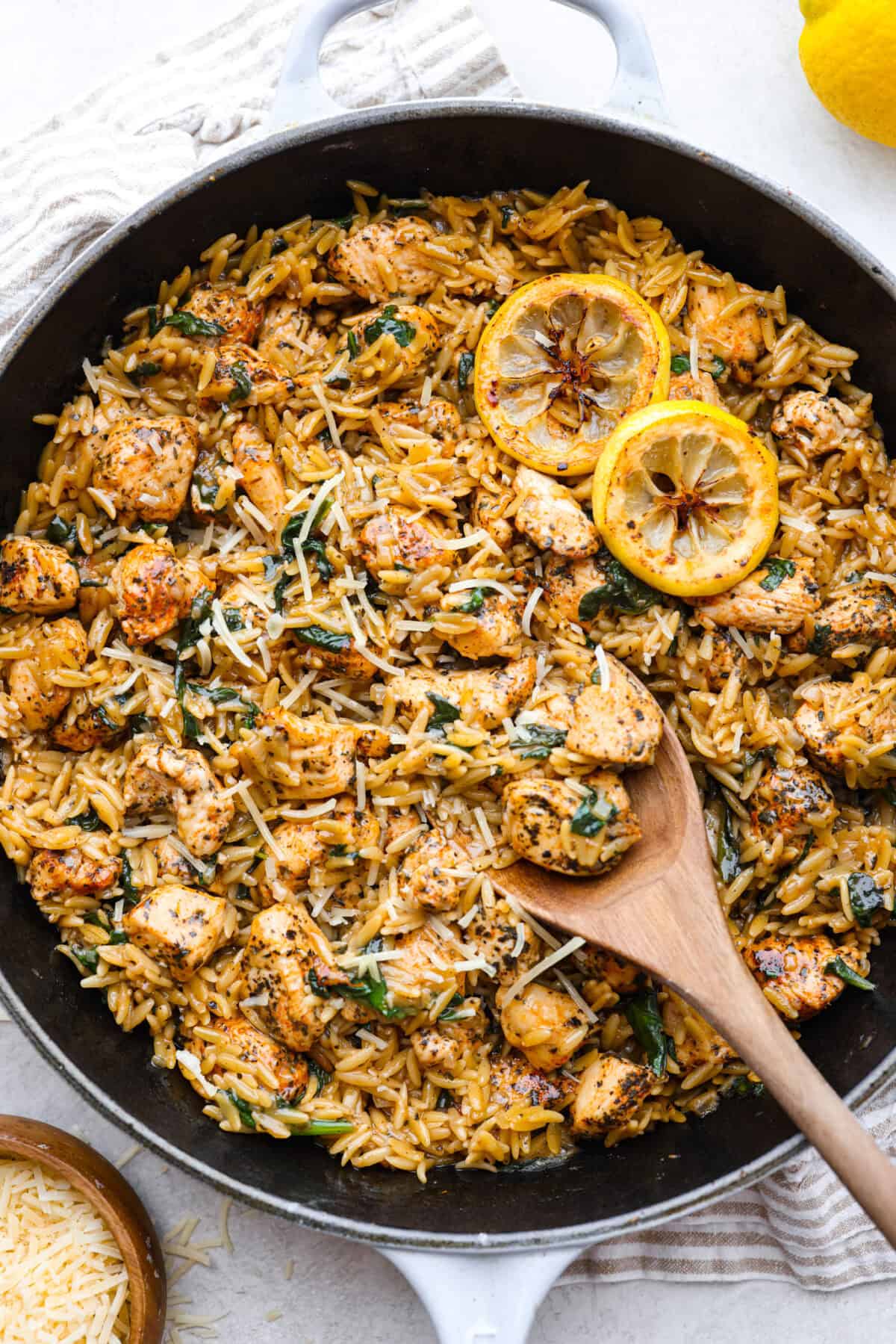 One-Pan Lemon Chicken Orzo is a comforting, well-rounded meal! Tender, juicy chicken pieces are joined with orzo pasta in a bright lemon sauce, resulting in melt-in-your-mouth deliciousness! Your whole family will adore this tasty dinner! Overhead shot of one-pan lemon chicken orzo, with a wooden spoon and lemon slices as garnish.