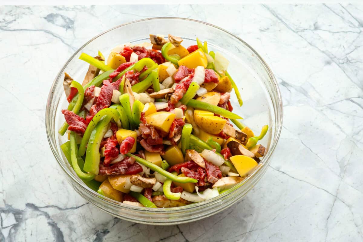 Try these delicious Philly Cheesesteak Foil Packs, a fun new way to enjoy the flavors of the classic Philly Cheesesteak. Tender beef, veggies, and melty cheese are grilled or baked together for a wonderful crowd-pleasing meal everyone will love! overhead shot of raw steak, peppers, onions, seasonings and potatoes in glass bowl.