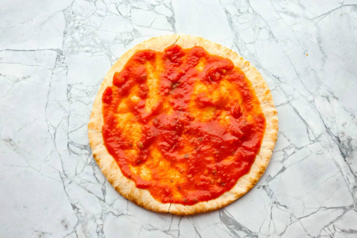 Pita pizza is like having your own personalized mini-pizza! Grab some ready-made pitas, pile on your favorite toppings, and indulge in a quick and tasty snack for a delightful way to satisfy your pizza craving whenever it strikes! Overhead shot of pita bread with marinara sauce spread out on it.