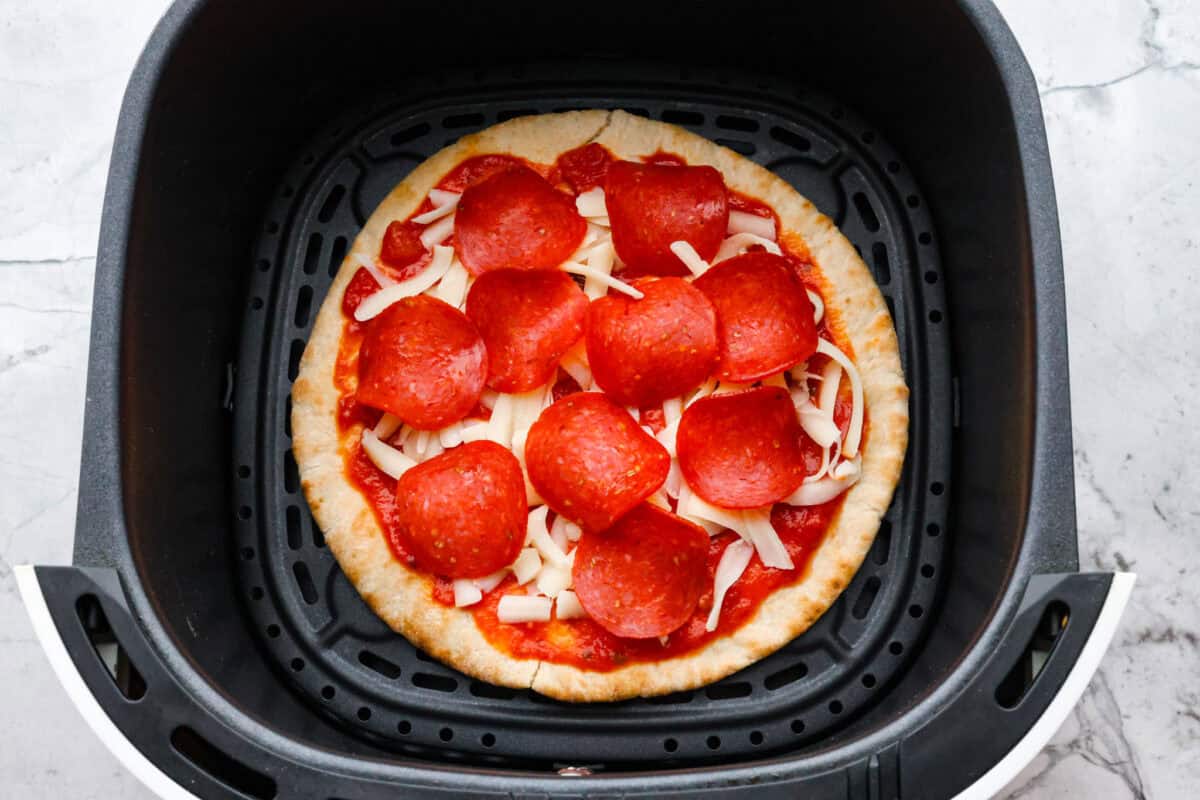 Pita pizza is like having your own personalized mini-pizza! Grab some ready-made pitas, pile on your favorite toppings, and indulge in a quick and tasty snack for a delightful way to satisfy your pizza craving whenever it strikes! Overhead shot of pita pizza at the bottom of air fryer basket ready to be cooked.