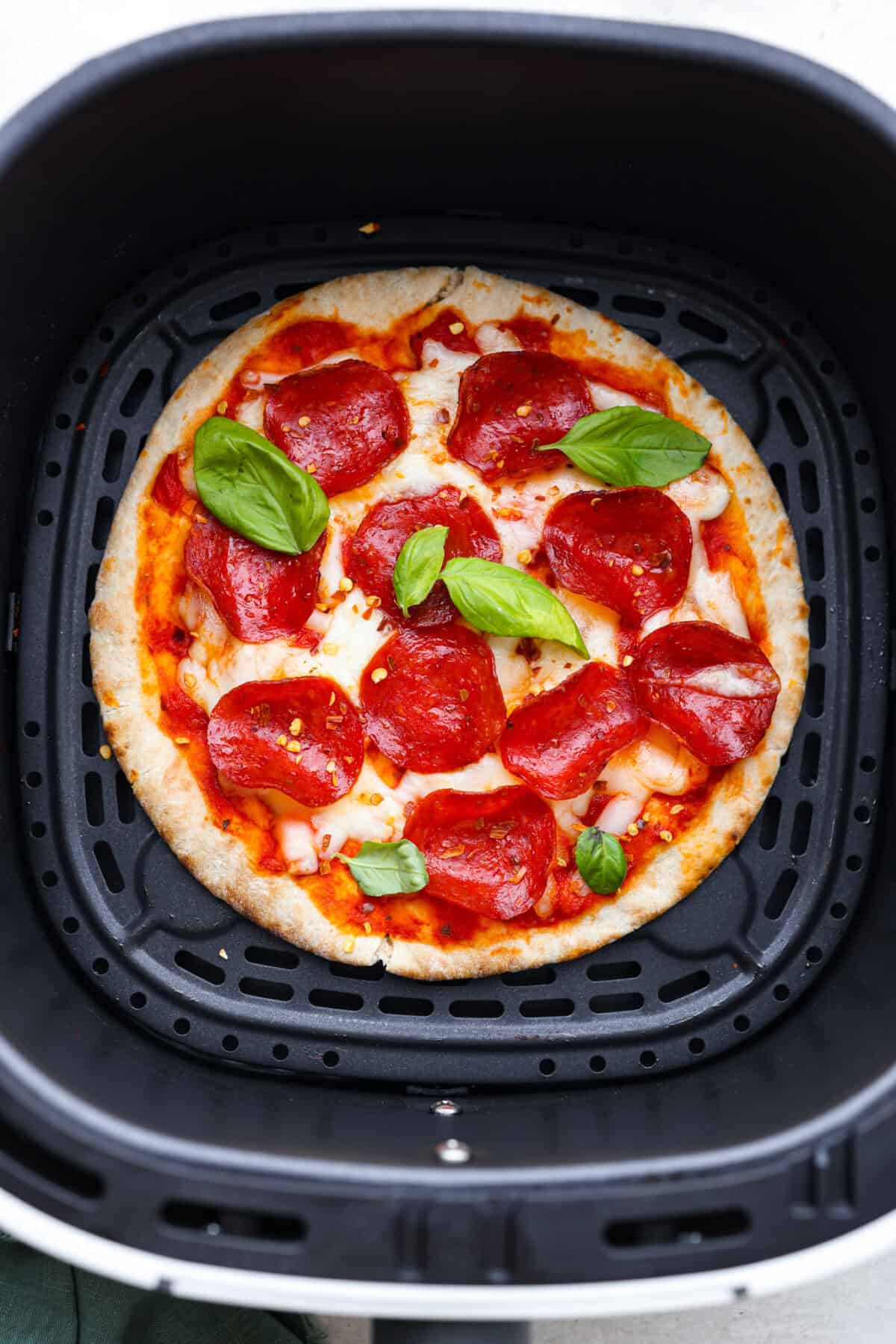 Pita pizza is like having your own personalized mini-pizza! Grab some ready-made pitas, pile on your favorite toppings, and indulge in a quick and tasty snack for a delightful way to satisfy your pizza craving whenever it strikes! Overhead shot of cooked pita pizza in the bottom of air fryer basket garnished with pepper flakes and basil.