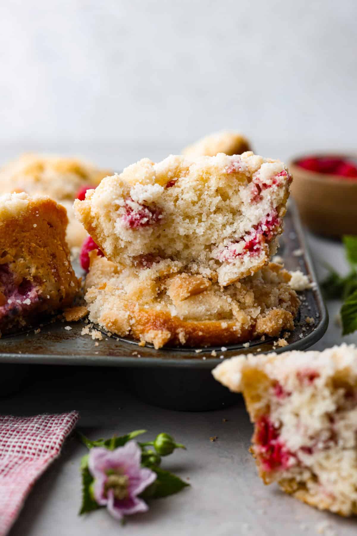 These raspberry muffins are moist and delicious, filled with plump fresh raspberries and topped with a sweet, crunchy streusel topping! They are perfect for a warm breakfast, brunch, or as an after-school or work snack. Your family will be begging for more, so donโt expect them to last long! Side shot of raspberry muffin cut in half sitting on top of a pan of raspberry muffins.