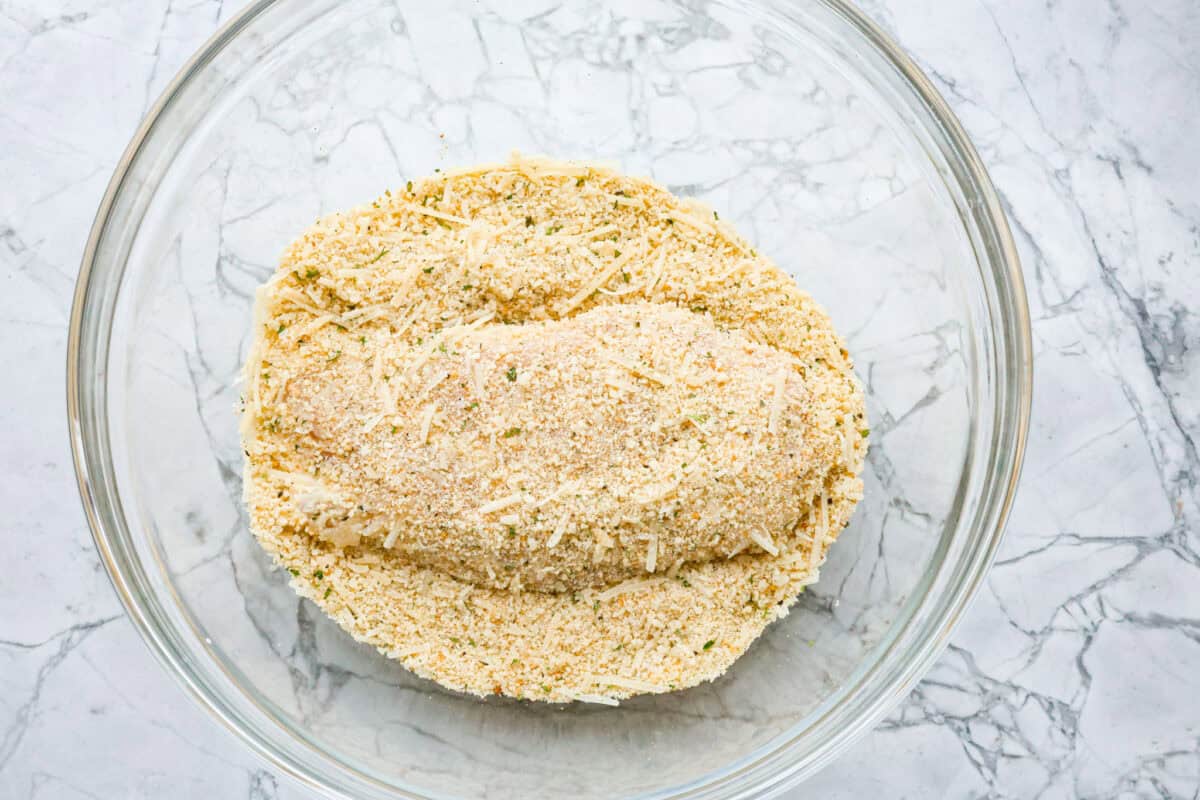 This simple slow-cooker chicken parmesan is full of delicious Italian flavors, oozy cheese, and mouthwatering marinara sauce. You'll be able to throw everything in the crock pot, set it, and forget it—dinner is served! Overhead shot of chicken in breading mixture in glass bowl.