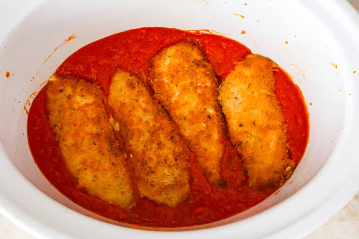 This simple slow-cooker chicken parmesan is full of delicious Italian flavors, oozy cheese, and mouthwatering marinara sauce. You'll be able to throw everything in the crock pot, set it, and forget it—dinner is served! Overhead shot of crockpot filled with marinara sauce and fried chicken cutlets on top of sauce.