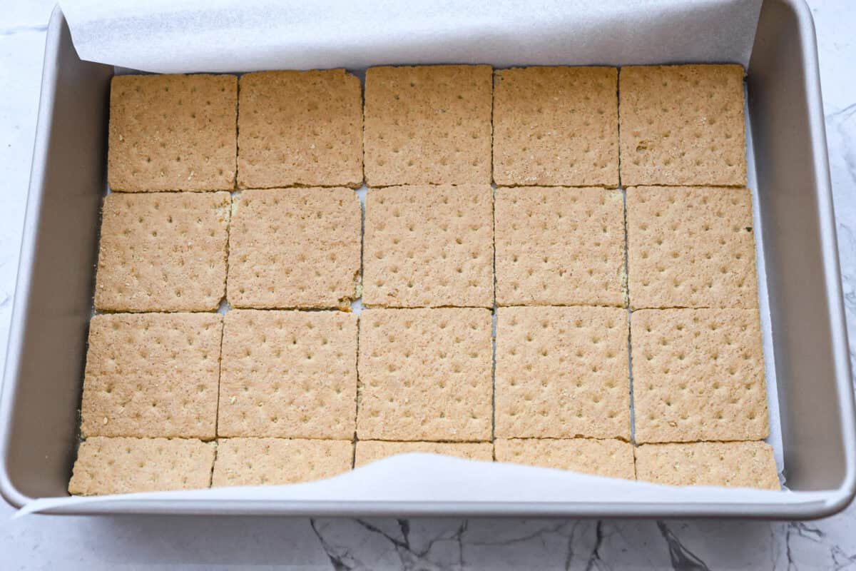 S'mores crack is a quick way to enjoy the classic s'more experience without a campfire. Featuring a buttery graham cracker crust, rich melted chocolate, and toasted marshmallows, this treat will quickly become a family favorite. Overhead shot of a pan with a layer of parchment covered in a single layer of graham crackers.
