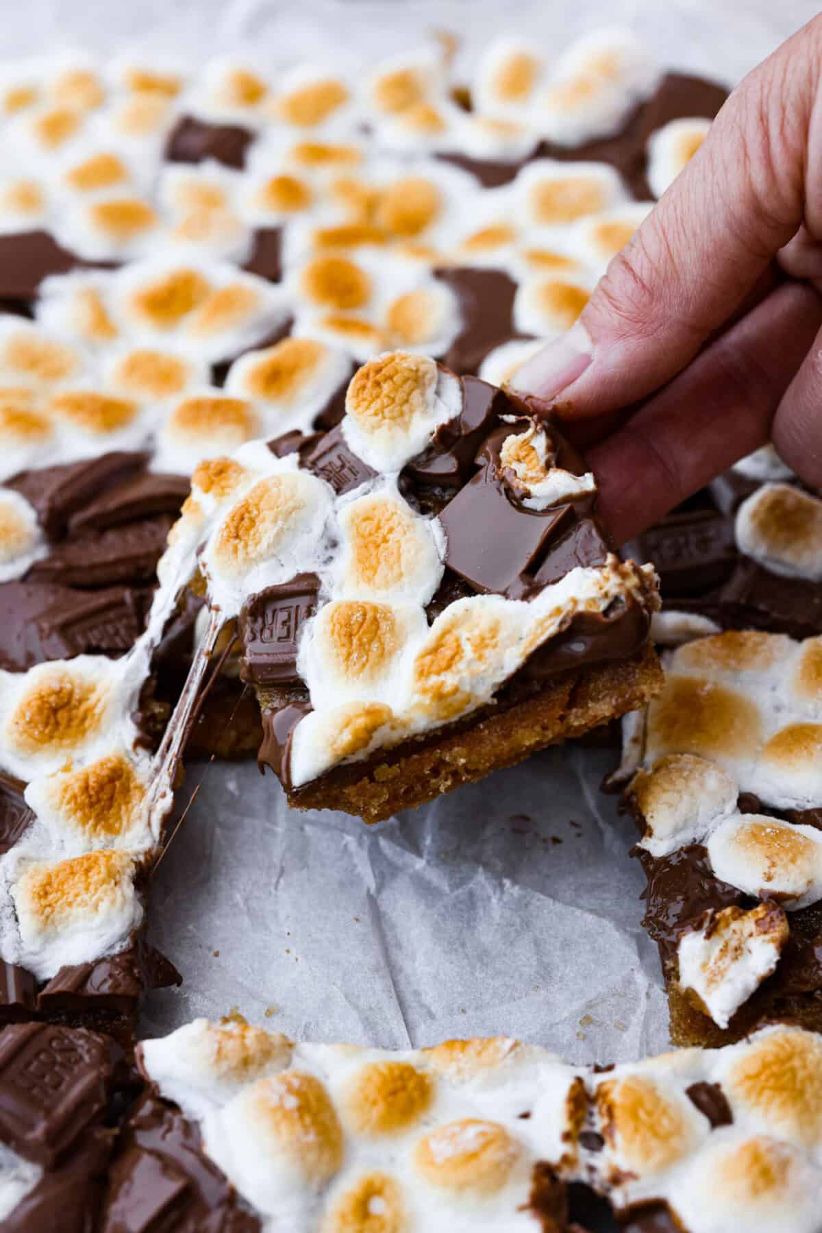 S'mores crack is a quick way to enjoy the classic s'more experience without a campfire. Featuring a buttery graham cracker crust, rich melted chocolate, and toasted marshmallows, this treat will quickly become a family favorite. Angled zoomed in shot of someone pulling a piece of gooey s'mores crack from the pan.