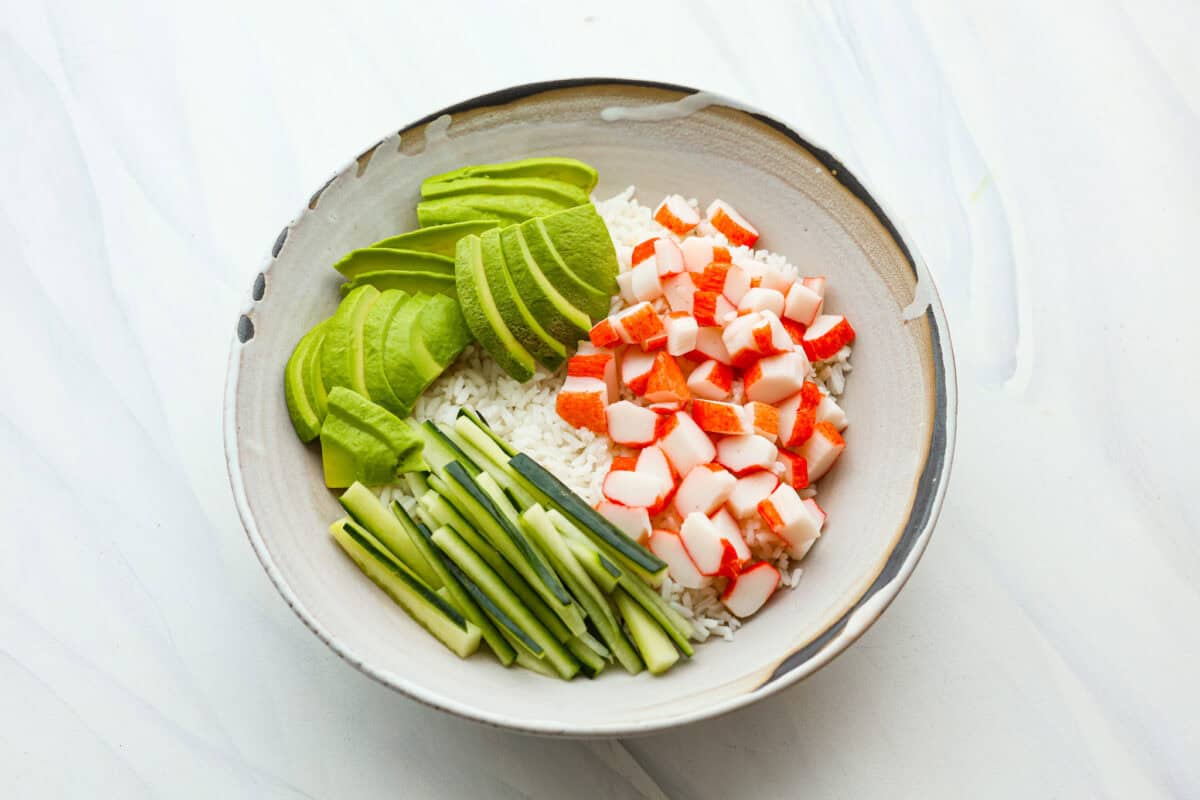 This sushi bowl is a deconstructed California sushi roll loaded with irresistible flavor and all of the best sushi ingredients. Itโs layered with sushi rice, crab, cucumber, avocado, a tasty sauce, and more. Itโs easy to make and ready to serve in just 20 minutes! Overhead shot of bowl with rice, crab, avocado and cucumber.