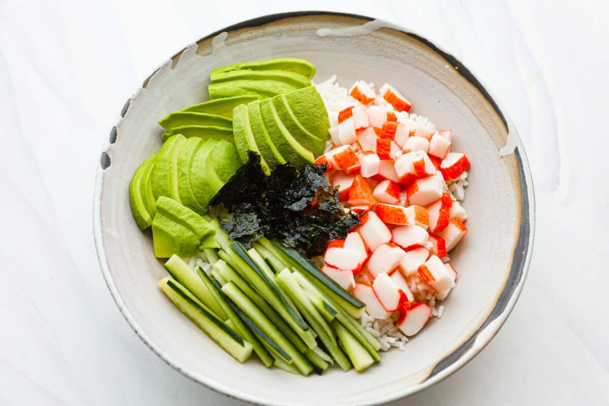 This sushi bowl is a deconstructed California sushi roll loaded with irresistible flavor and all of the best sushi ingredients. Itโs layered with sushi rice, crab, cucumber, avocado, a tasty sauce, and more. Itโs easy to make and ready to serve in just 20 minutes! Overhead shot of bowl with rice, crab, avocado, cucumber and seaweed.