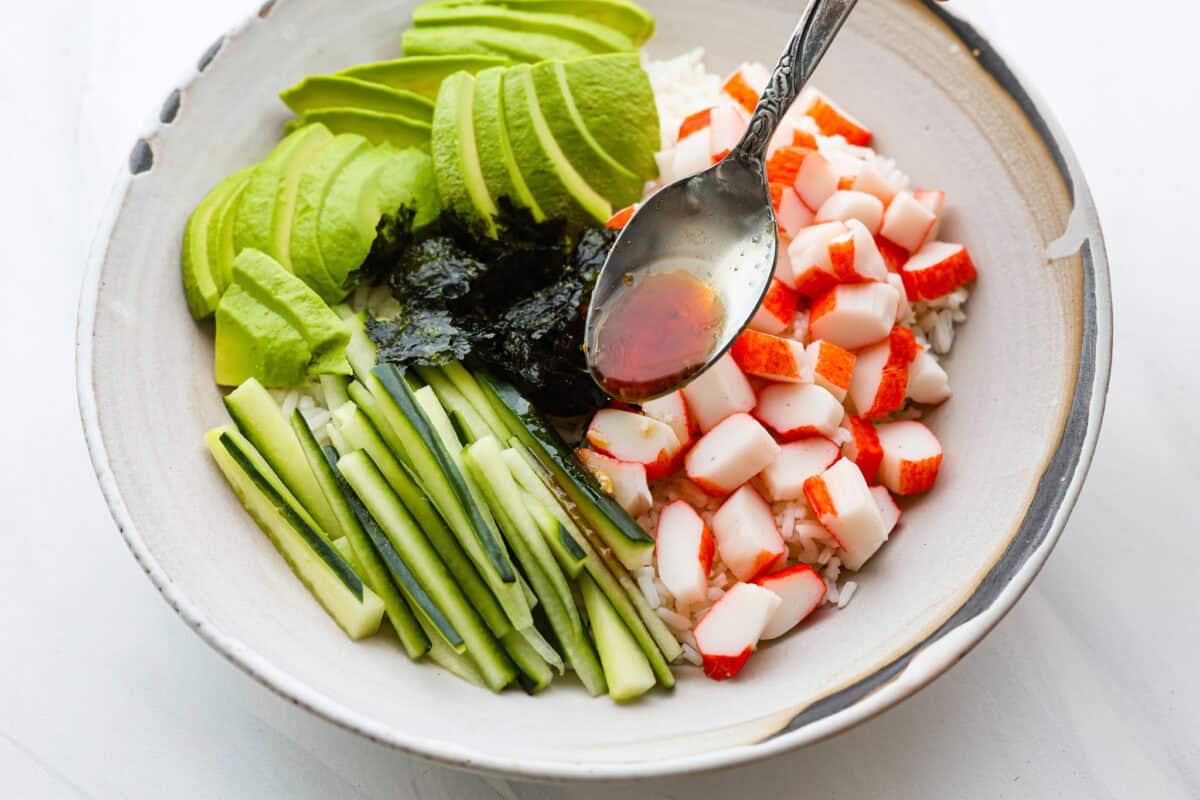 This sushi bowl is a deconstructed California sushi roll loaded with irresistible flavor and all of the best sushi ingredients. Itโs layered with sushi rice, crab, cucumber, avocado, a tasty sauce, and more. Itโs easy to make and ready to serve in just 20 minutes! Overhead shot of sushi bowl with someone pouring sauce over it with a spoon.