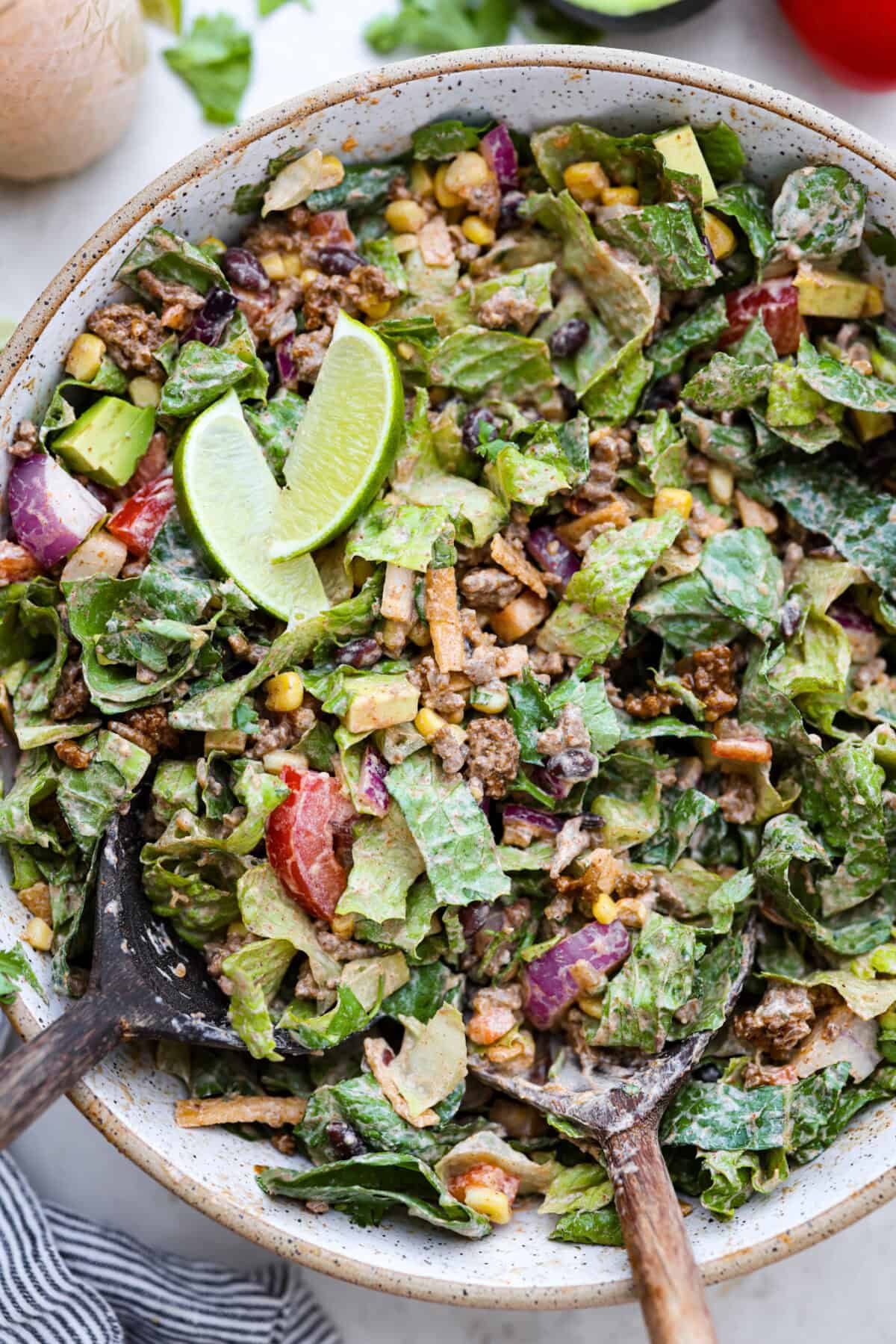 Beef taco salad is a delicious and simple family favorite, perfect for summer evenings! Packed with your favorite taco essentials like beans, corn, avocado, and fresh cilantro, it's bursting with flavor and sure to become a staple dish! Overhead shot of completed beef taco salad in large bowl, with limes wedges on top.
