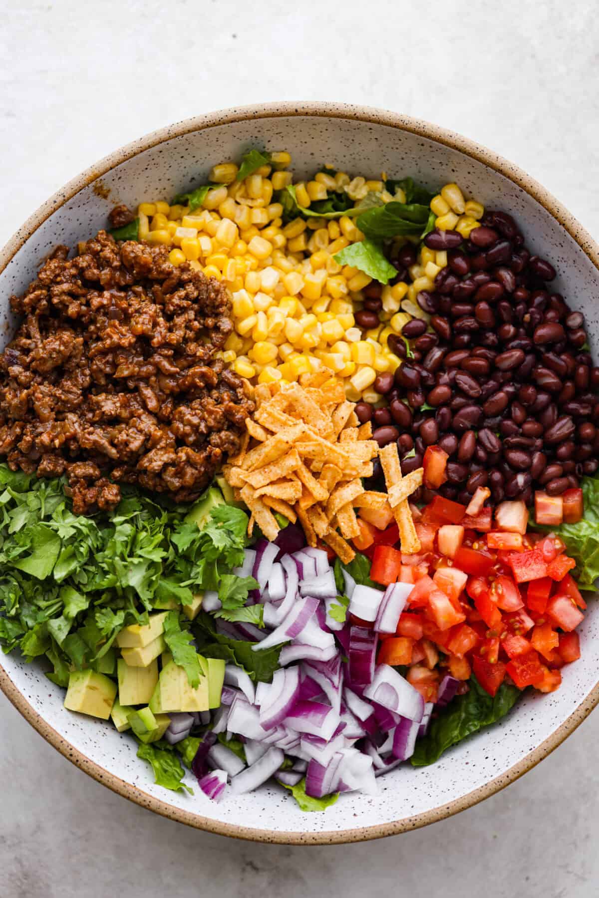 Beef taco salad is a delicious and simple family favorite, perfect for summer evenings! Packed with your favorite taco essentials like beans, corn, avocado, and fresh cilantro, it's bursting with flavor and sure to become a staple dish! Overhead shot of separated salad ingredients in large bowl.