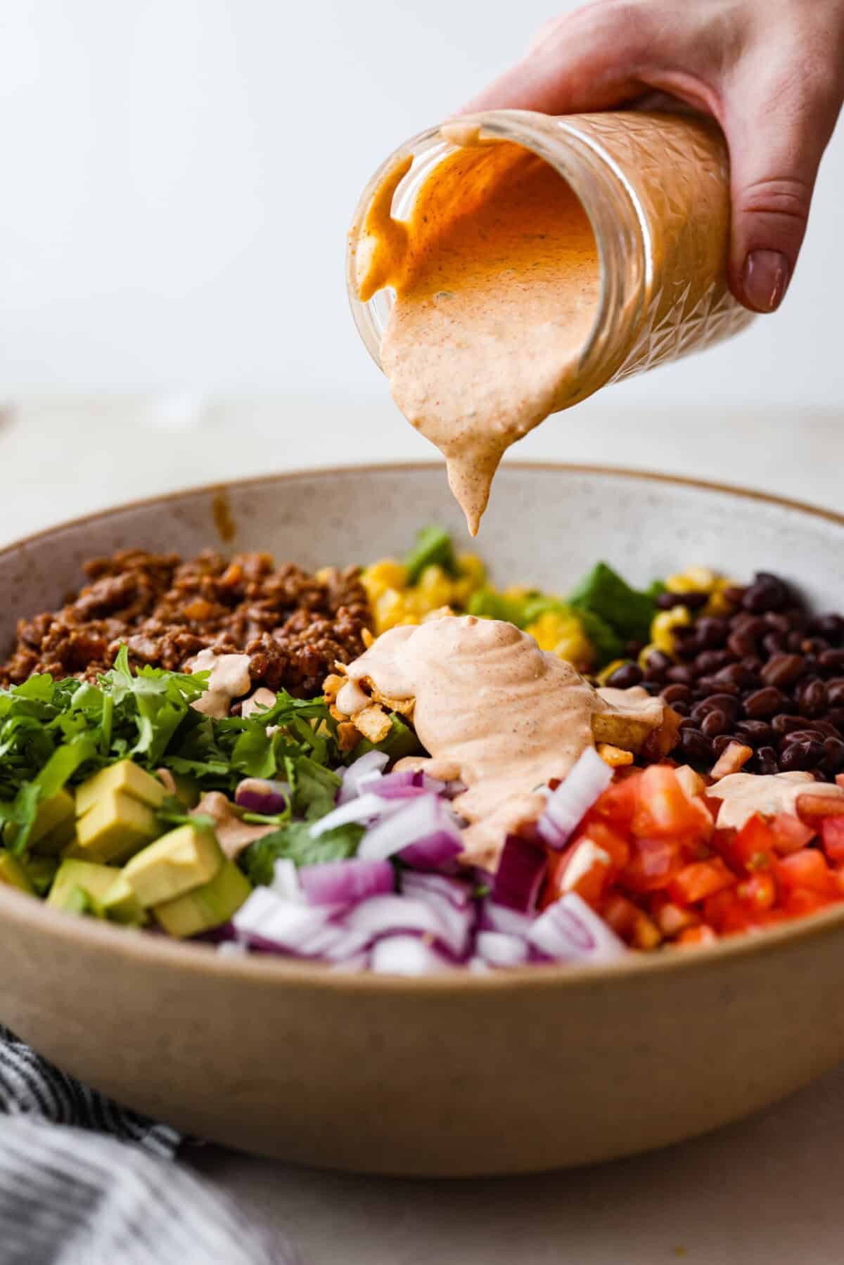 Beef taco salad is a delicious and simple family favorite, perfect for summer evenings! Packed with your favorite taco essentials like beans, corn, avocado, and fresh cilantro, it's bursting with flavor and sure to become a staple dish! Side shot of someone pouring salad dressing of the taco salad.