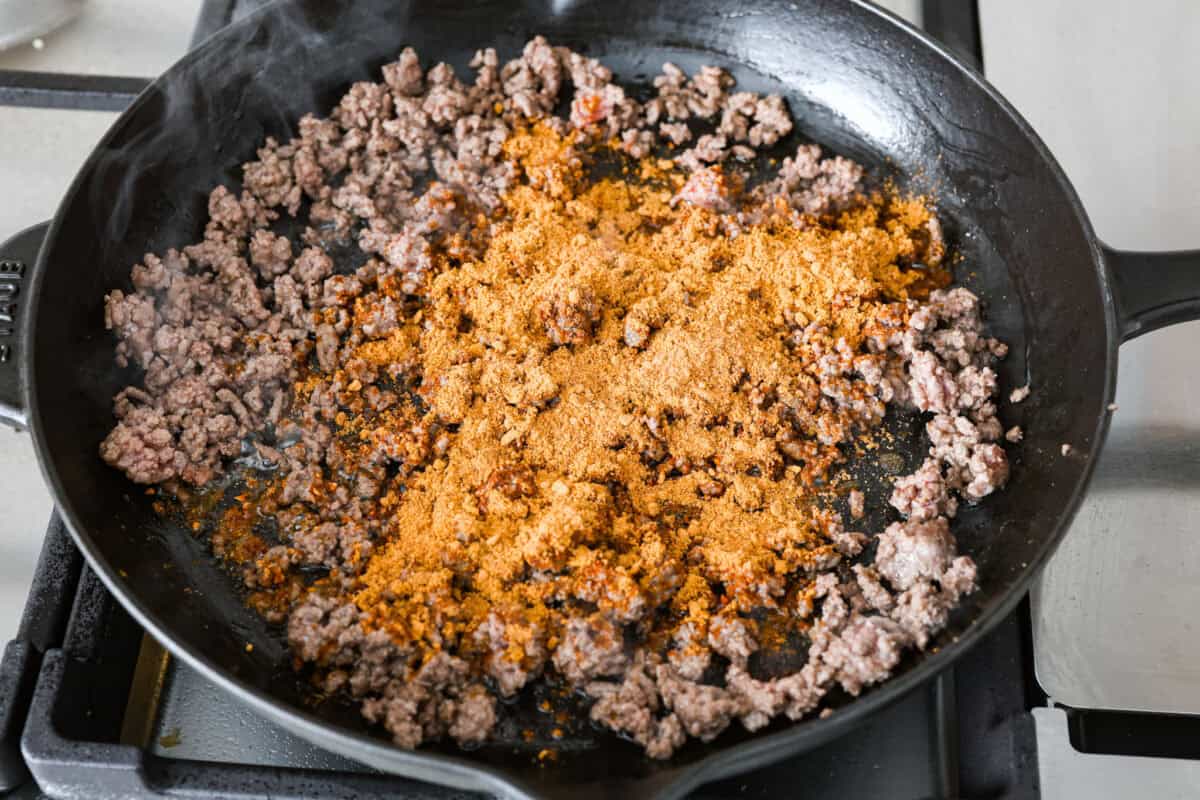 Tater Tot Nachos (Totchos) are made with crispy golden tater tots, topped with seasoned beef, scoops of refried beans, corn, black beans, salsa and melty cheese. Add your favorite toppings and dig in! Overhead shot of ground beef being cooked in a skillet with taco seasoning.