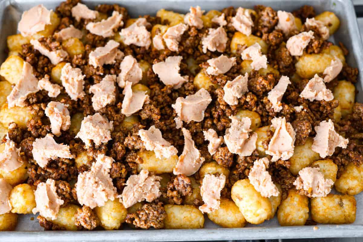 Tater Tot Nachos (Totchos) are made with crispy golden tater tots, topped with seasoned beef, scoops of refried beans, corn, black beans, salsa and melty cheese. Add your favorite toppings and dig in! overhead shot of prepped pan full of tater tots, seasoned ground beef, and dollops of refried beans.