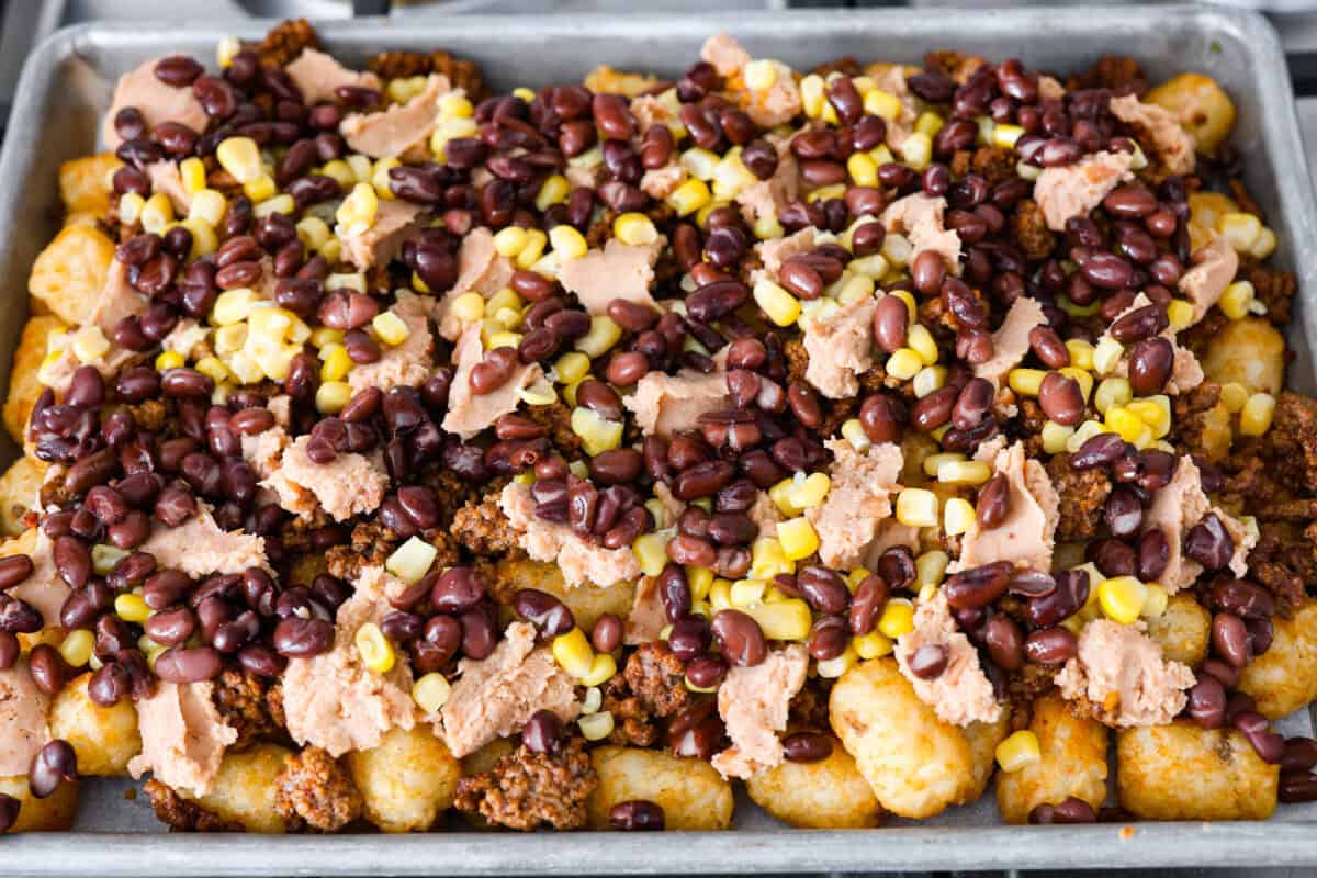 Tater Tot Nachos (Totchos) are made with crispy golden tater tots, topped with seasoned beef, scoops of refried beans, corn, black beans, salsa and melty cheese. Add your favorite toppings and dig in! Overhead shot of prepared pan with added black beans and corn.