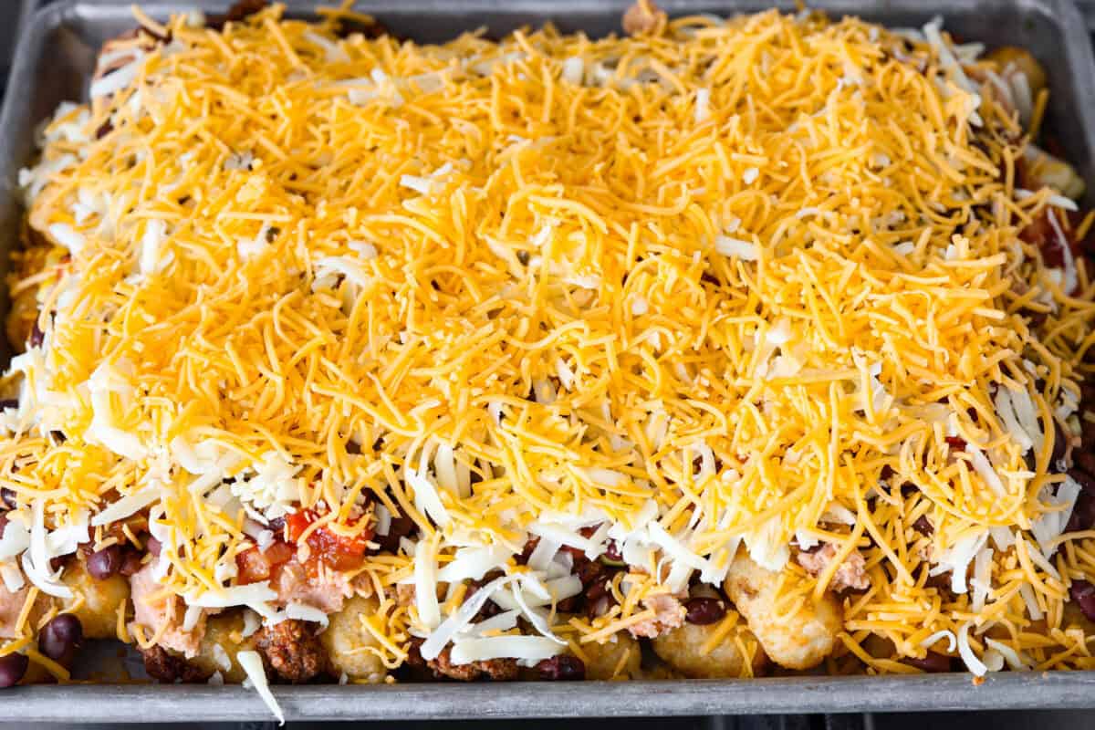 Tater Tot Nachos (Totchos) are made with crispy golden tater tots, topped with seasoned beef, scoops of refried beans, corn, black beans, salsa and melty cheese. Add your favorite toppings and dig in! Overhead shot of prepared pan of tater tot nachos with shredded cheese over the top.