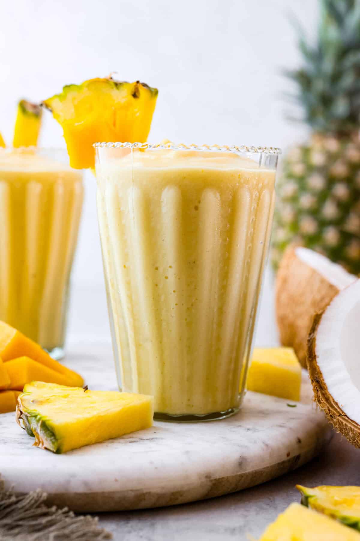 This refreshing tropical smoothie is bursting with tropical fruit flavors, making it the perfect treat for a hot day. Sip it, and you'll feel like you're lounging on the beach, enjoying a drink freshly served by the cabana boy! Side shot of glass of tropical smoothie with wedge of pineapple on the rim of the glass.