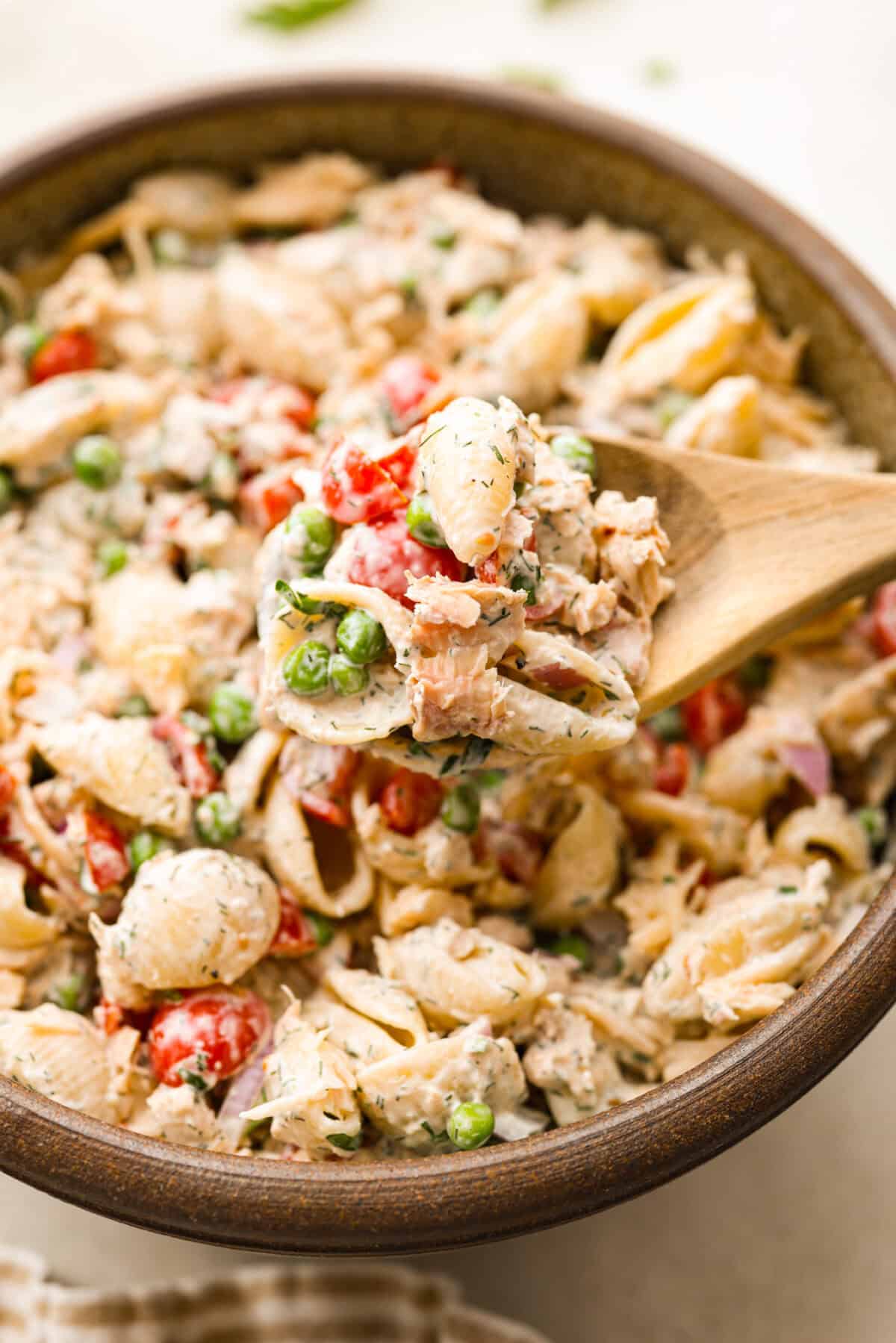 Tuna pasta salad is a perfect protein-packed lunch or side dish! It combines tuna, veggies, and pasta then gets tossed in a creamy dill dressing. Close side view of a wood spoon lifted a scoop of tuna pasta salad.