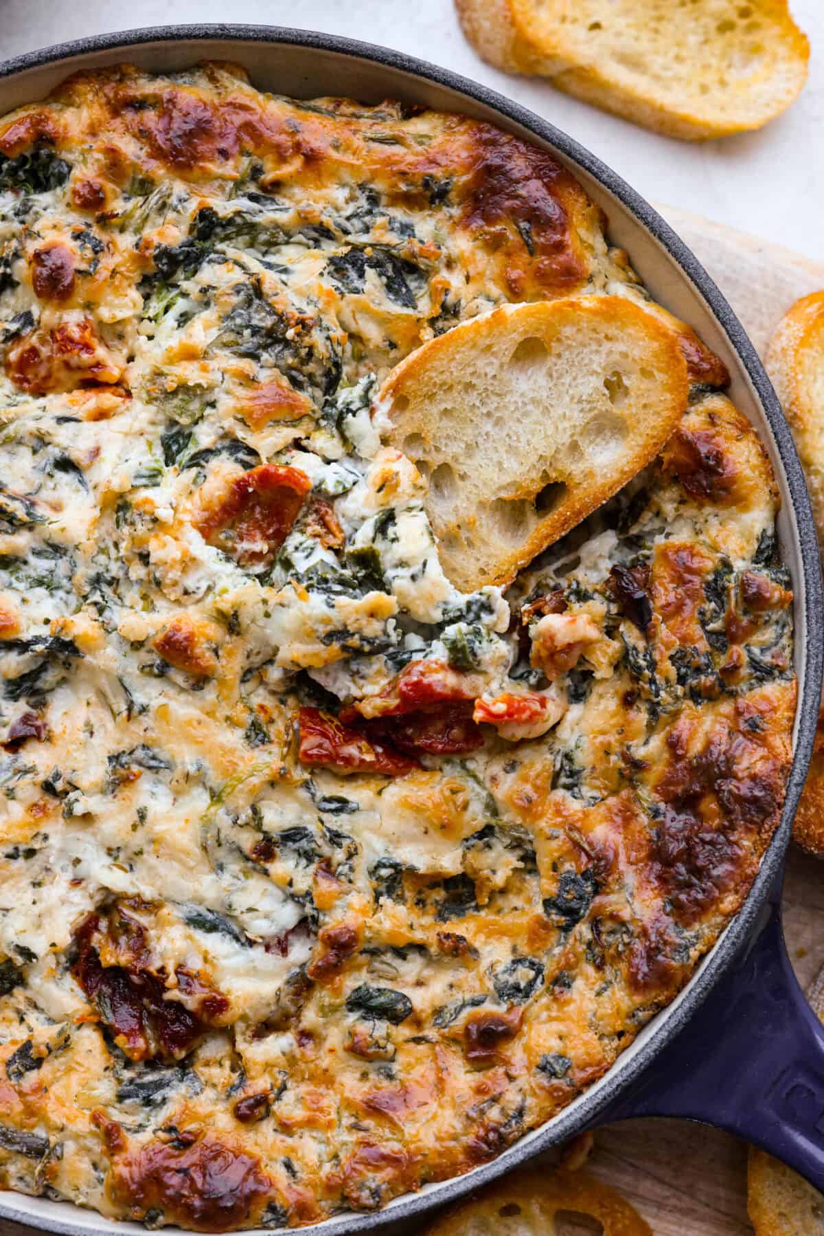 There's a new appetizer in town, and this creamy Tuscan dip won everyone's hearts! The combination of cheeses, garlic, spinach, and sun-dried tomatoes creates an addicting dip you’ll want to bring to all of your gatherings! Close top view of creamy Tuscan dip in a blue skillet with a crostini dipping in.