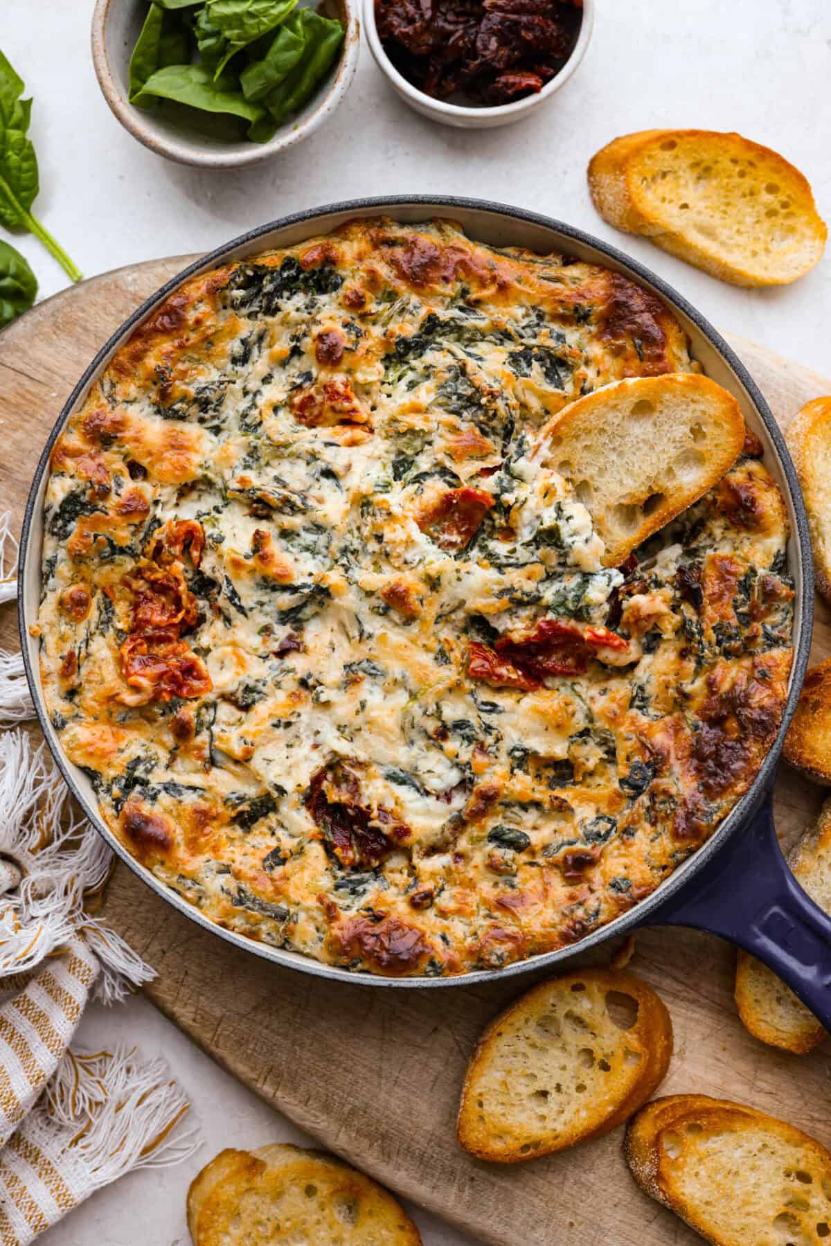 There's a new appetizer in town, and this creamy Tuscan dip won everyone's hearts! The combination of cheeses, garlic, spinach, and sun-dried tomatoes creates an addicting dip you’ll want to bring to all of your gatherings! Top view of creamy Tuscan dip in a skillet with crostini scattered on a wood board.