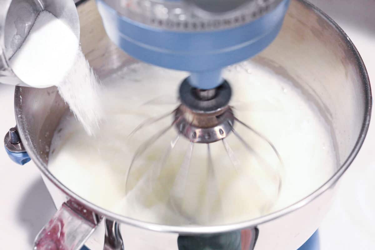 This light & fluffy Angel Food Cake is like a slice of heaven on a plate! It's delicious all on its own but absolutely divine topped with sweetened whipped cream and fresh berries or lemon curd! Overhead shot of sifted sugar being poured into a stand mixer whisking egg whites.