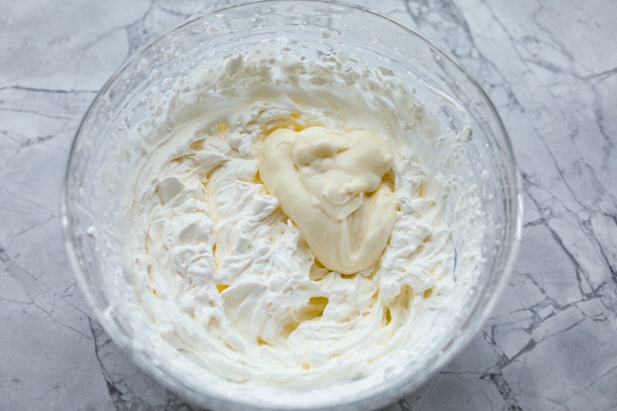 Rich and creamy Cinnamon Roll Cheesecake has the delicious flavor of a cinnamon roll in decadent cheesecake form. This irresistible dessert is made with a cinnamon graham cracker crust, a velvety cream cheese filling with spiced cinnamon throughout, and a classic whipped cream topping. Overhead shot of a dollop of cream cheese mixture in the whipped cream in glass bowl.
