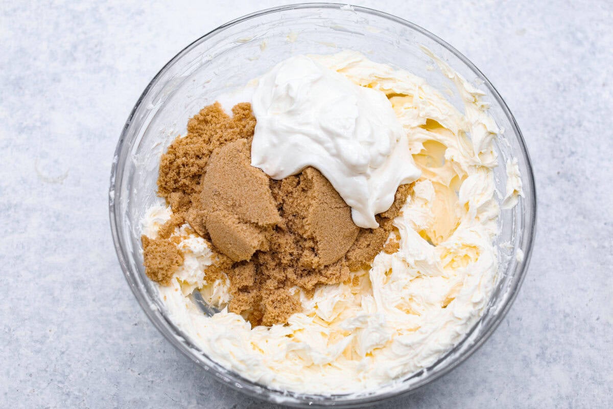 Rich and creamy Cinnamon Roll Cheesecake has the delicious flavor of a cinnamon roll in decadent cheesecake form. This irresistible dessert is made with a cinnamon graham cracker crust, a velvety cream cheese filling with spiced cinnamon throughout, and a classic whipped cream topping. Overhead shot of brown sugar, cream cheese, and sour cream in glass bowl.