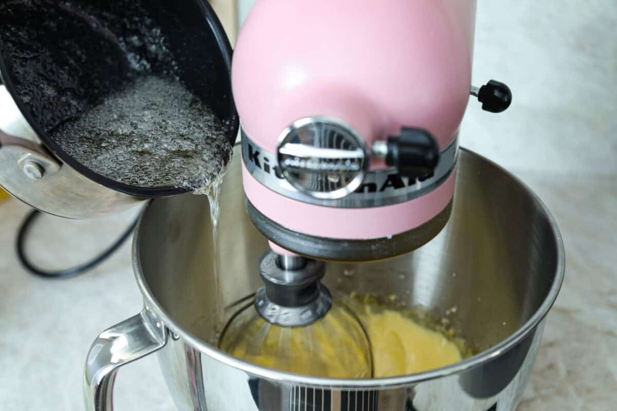 French buttercream is a luxurious, creamy frosting that will wow your guests and tantalize your taste buds. The elegant flavor comes from using egg yolks as the base and delicious, simple syrup for sweetness. Elevate your baking game with this unique recipe. Angle shot of sugar syrup being slowly poured into a stand mixer with whipped egg whites.