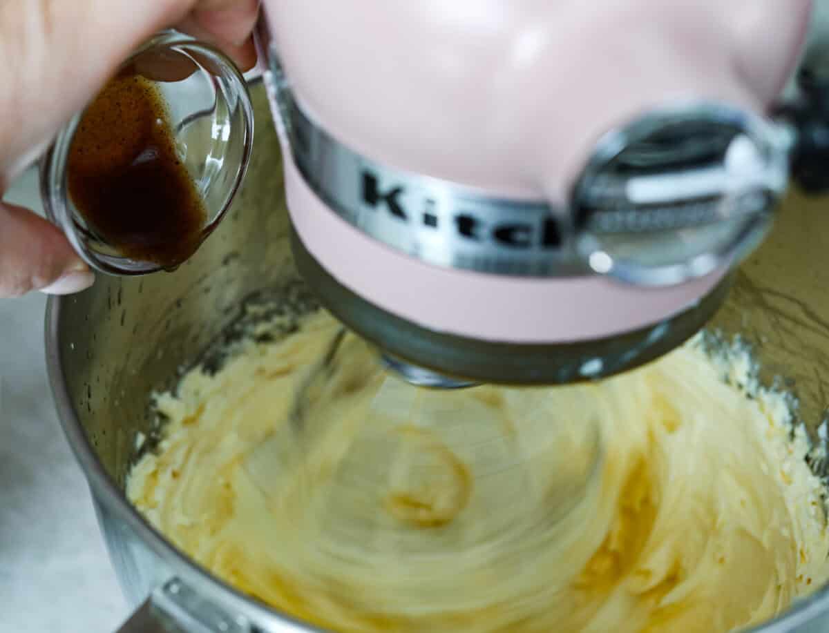 French buttercream is a luxurious, creamy frosting that will wow your guests and tantalize your taste buds. The elegant flavor comes from using egg yolks as the base and delicious, simple syrup for sweetness. Elevate your baking game with this unique recipe. Vanilla being poured into stand mixer bowl whipping frosting.