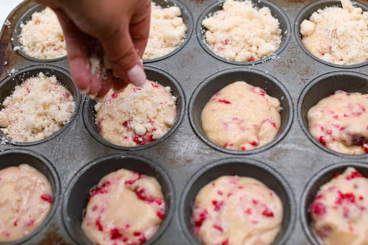 These raspberry muffins are moist and delicious, filled with plump fresh raspberries and topped with a sweet, crunchy streusel topping! They are perfect for a warm breakfast, brunch, or as an after-school or work snack. Your family will be begging for more, so donโt expect them to last long! Angle shot of someone sprinkling streusel over muffin batter in muffin tin.
